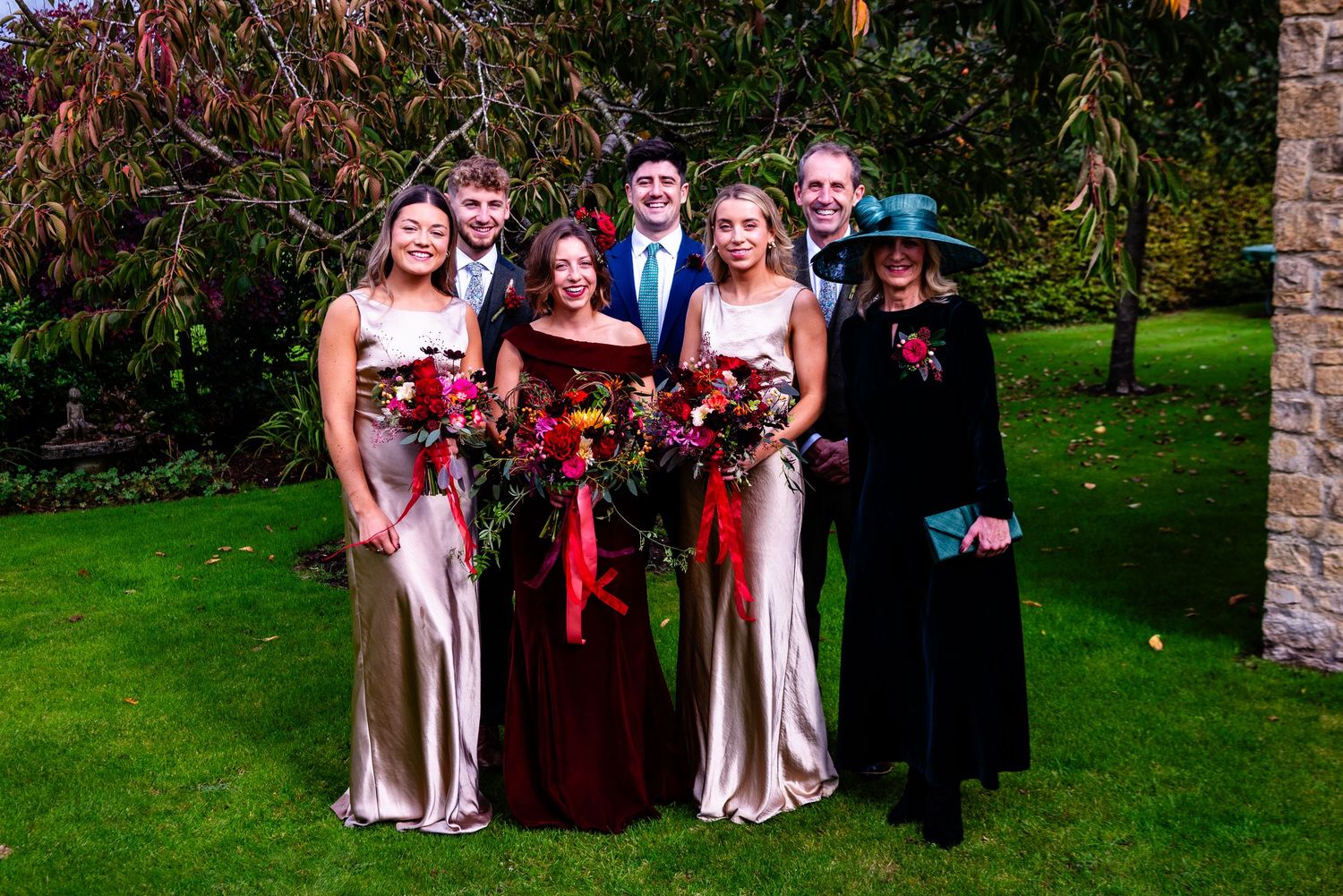 Wedding party posing outdoors on green lawn, bridesmaids in gold and burgundy gowns holding vibrant red bouquets.