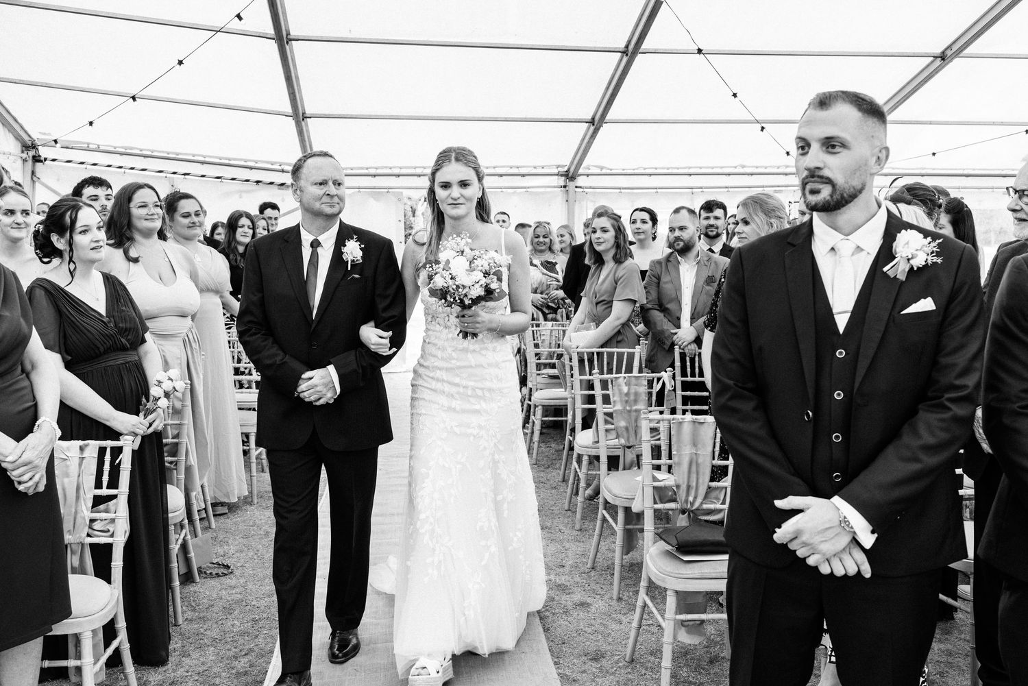 Black and white wedding photo of bride walking down the aisle with her father toward the waiting groom.