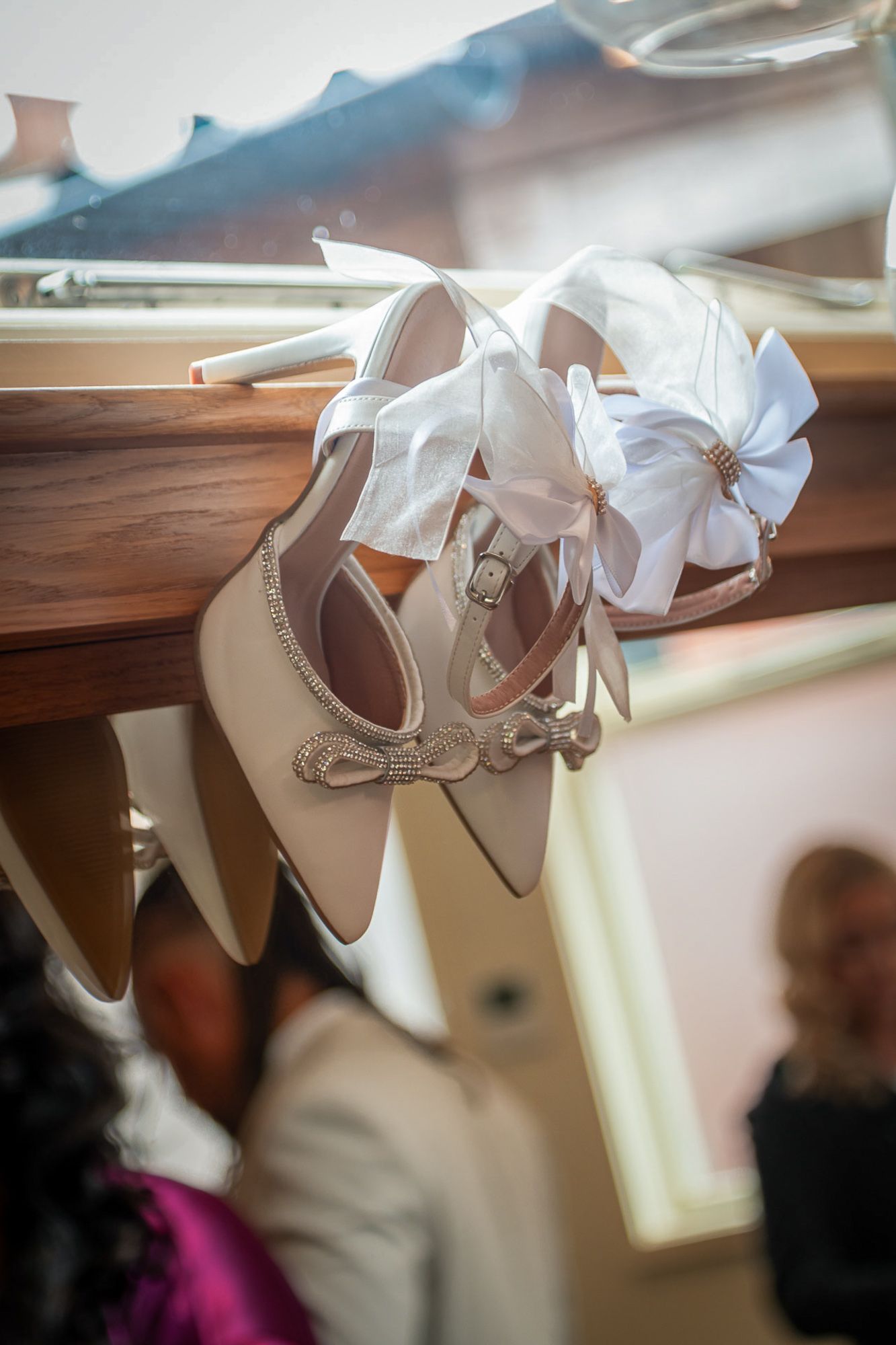 Elegant white bridal heels with satin bow and crystal embellishments hanging on a wooden rail in soft natural light.