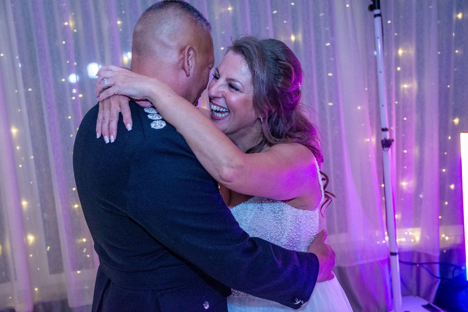 Bride and groom share a joyful first dance embrace under twinkling fairy lights at wedding reception.