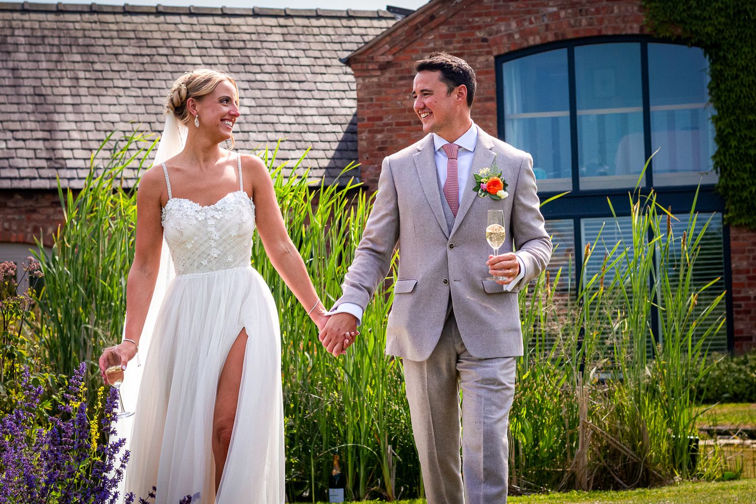 Smiling bride and groom walk hand in hand outside a brick venue on a sunny wedding day.