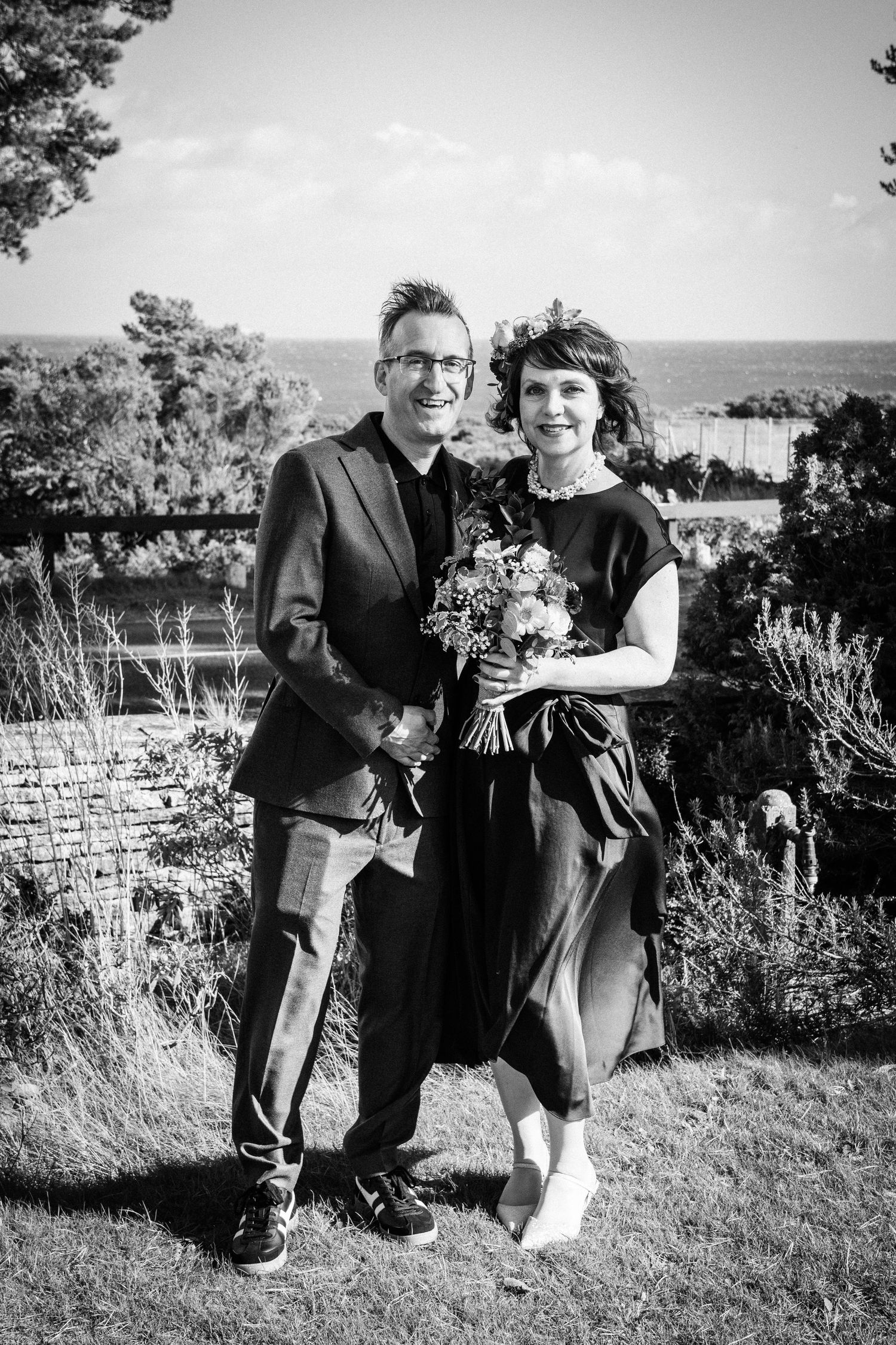 Black and white photo of smiling couple standing outdoors with ocean view, bride holding wildflower bouquet.