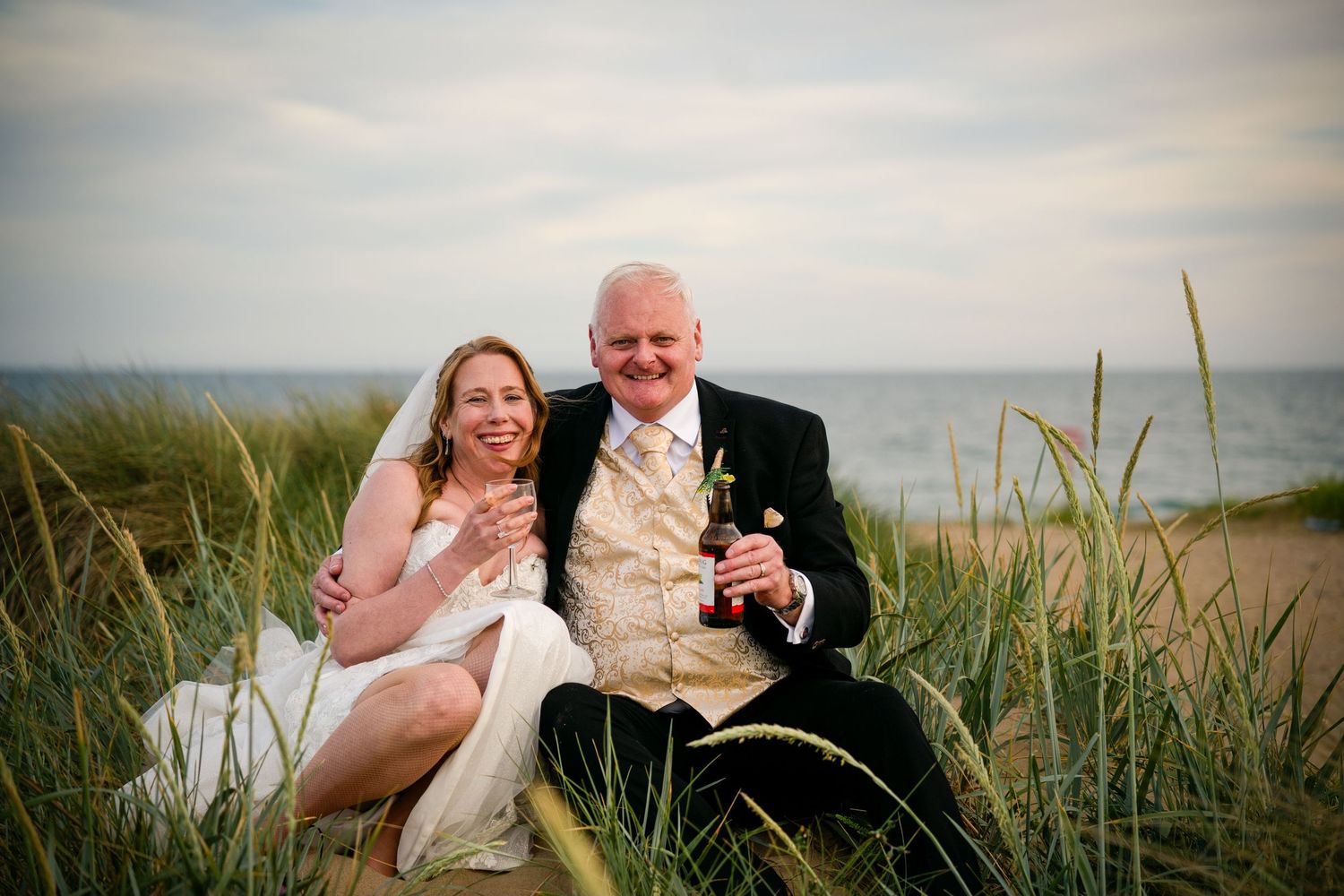 Happy bride and groom sitting together in beach grass by the ocean, smiling and holding drinks on their wedding day.