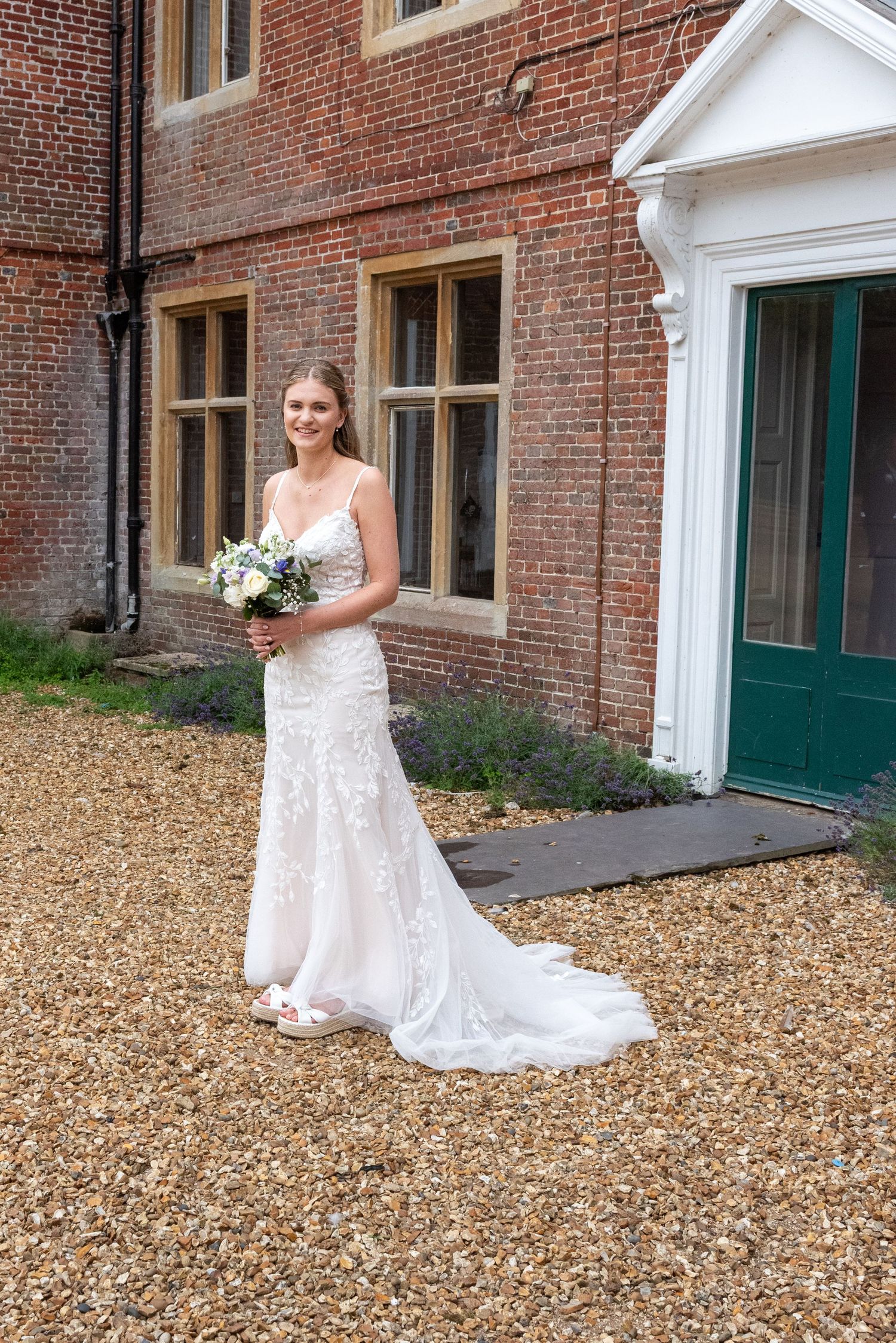 Bride in a lace wedding gown holding a bouquet outside a classic red brick manor house.