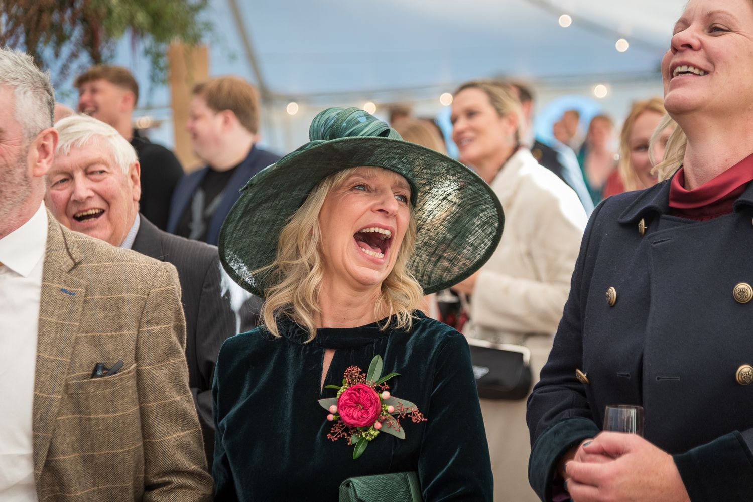 Woman in green hat laughing joyfully at a wedding reception surrounded by guests.