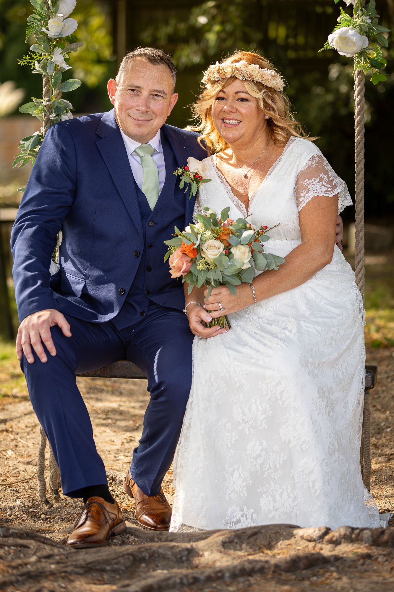 Bride and groom sitting on a rope swing outdoors, bride holding floral bouquet wearing lace dress and flower crown.