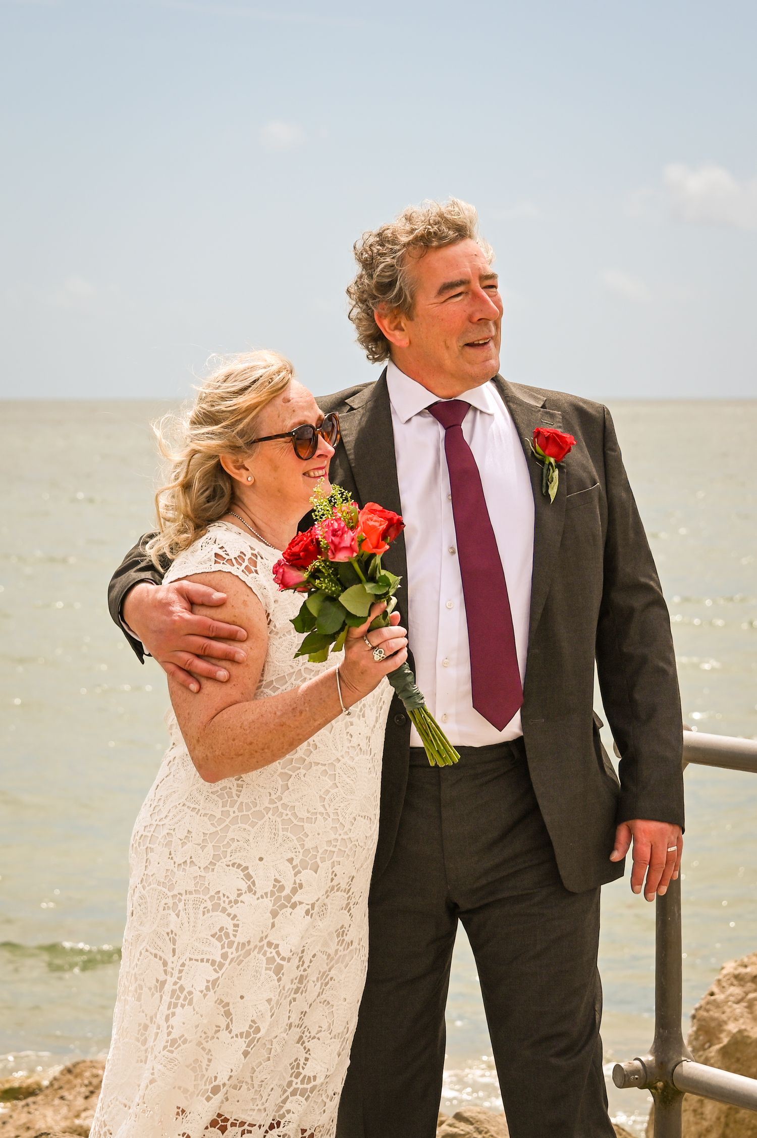 Older couple in wedding attire holding red roses by the sea, bride in lace dress with sunglasses, groom in suit with burgundy tie.