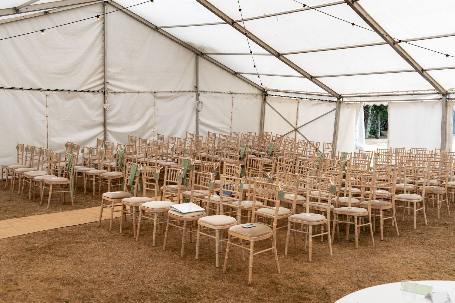 Rows of elegant wooden Chiavari chairs with cream cushions arranged in a spacious white marquee tent for a wedding ceremony.