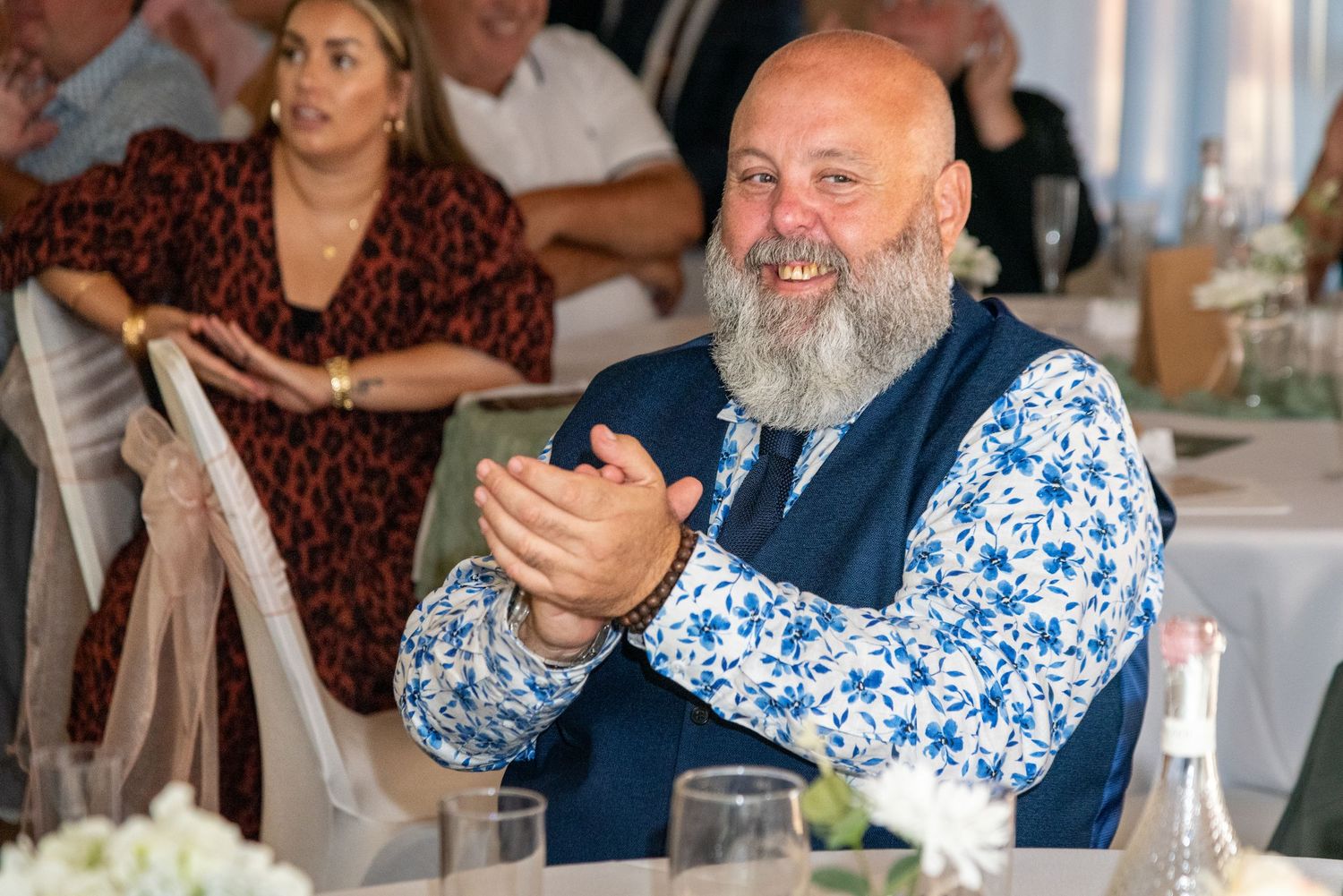 Bald man with gray beard clapping and smiling at a wedding reception, wearing a blue floral shirt and navy vest.