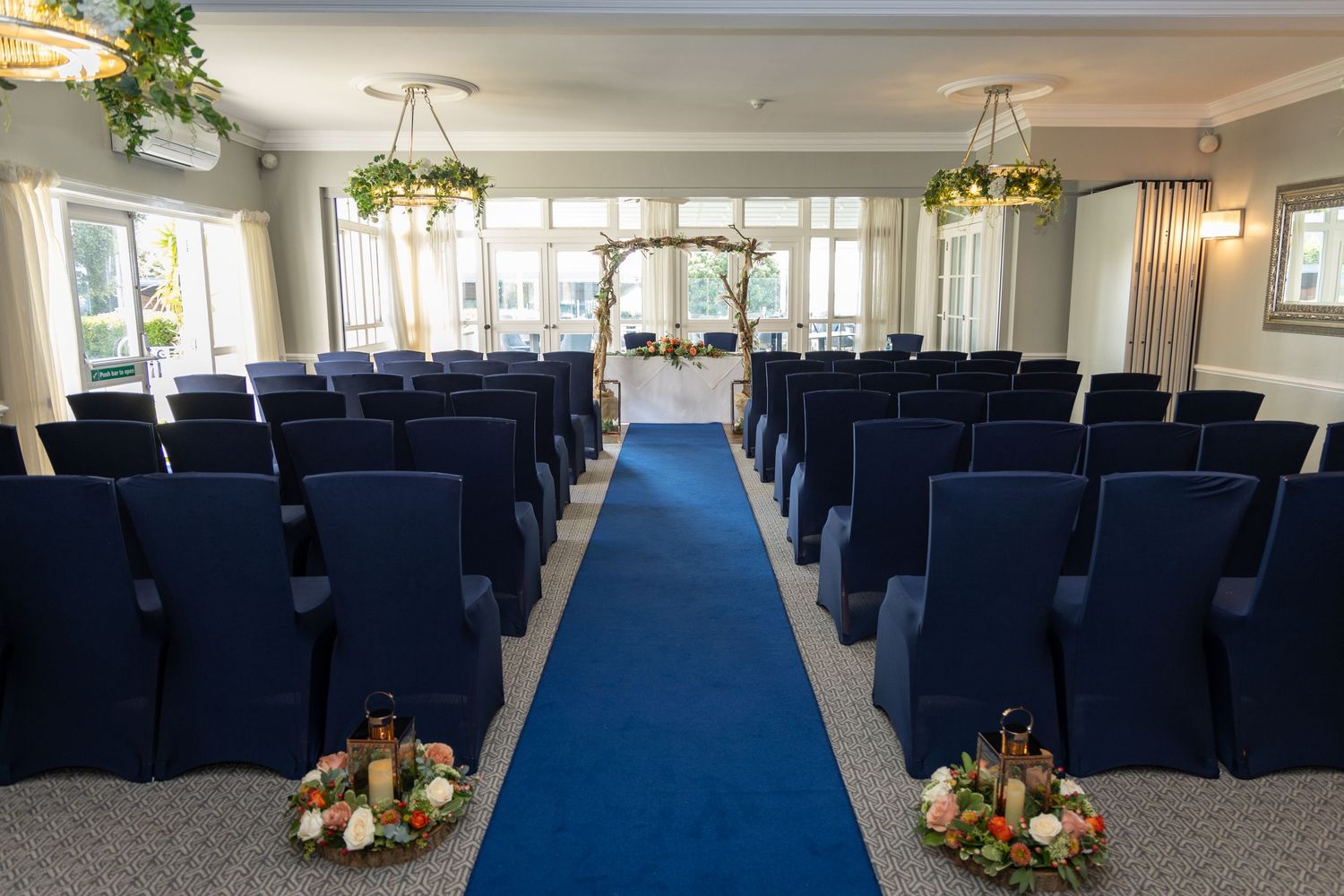 Indoor wedding ceremony venue with blue carpet aisle, navy chairs, floral arch and hanging greenery decorations.