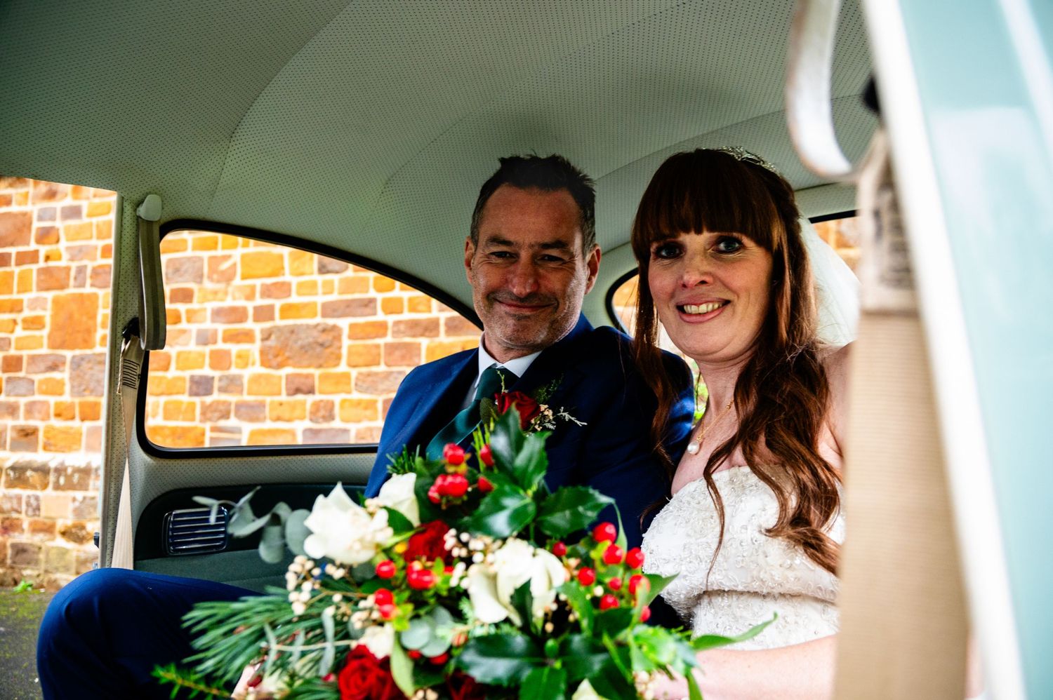 A couple smiles in a vintage car with wedding flowers and decorations visible through the window.