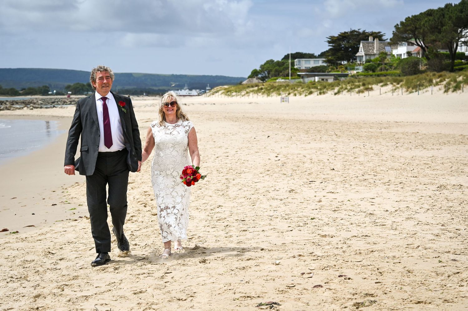 Couple in wedding attire walking hand-in-hand on a sandy beach, bride holding red bouquet.