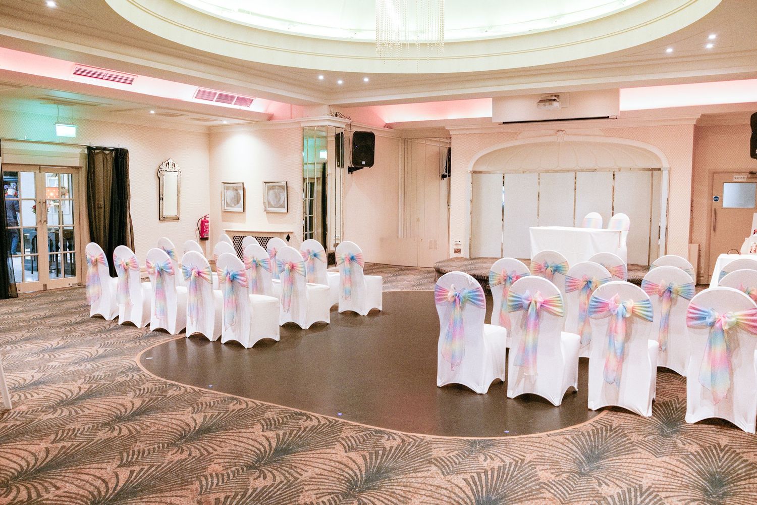 Elegant wedding venue with white chair covers, pink sashes, and soft lighting arranged in a semicircle on a dance floor.