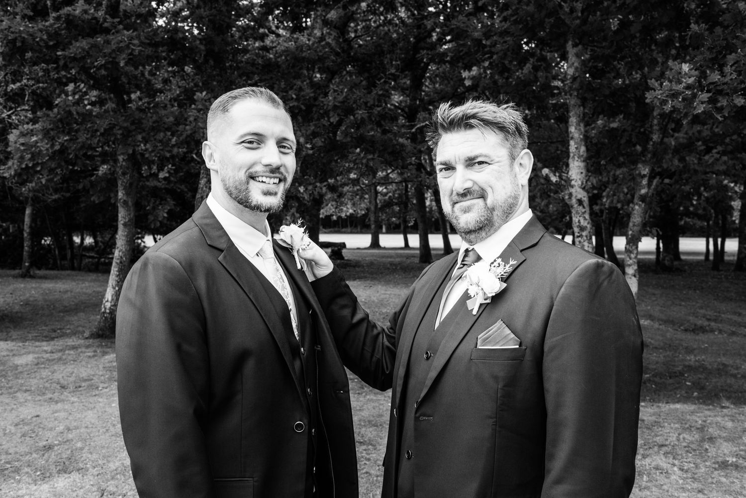 Two grooms in suits pose outdoors among trees in a classic black and white wedding portrait photo.