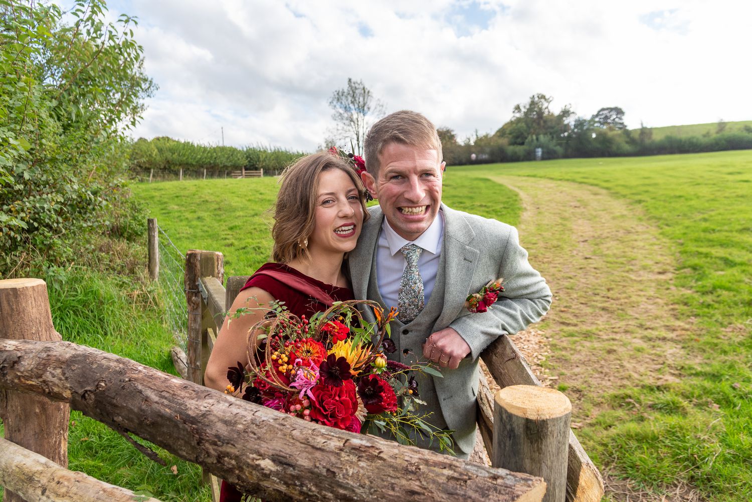 Happy couple posing by a rustic wooden fence in a green countryside field, bride holding a vibrant red bouquet.