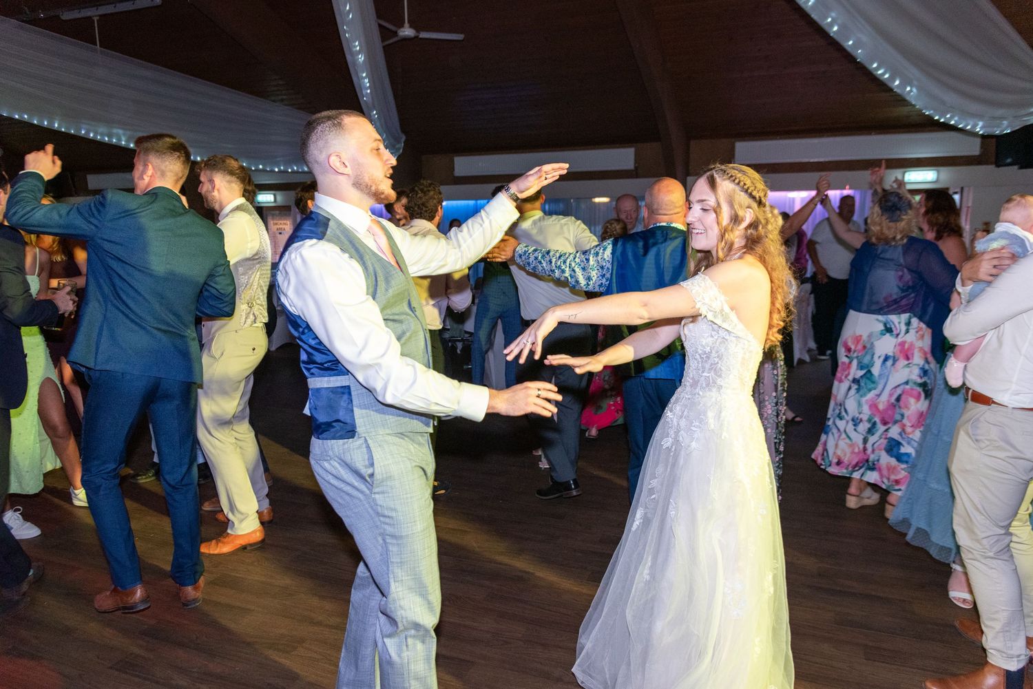 Bride and groom dancing joyfully at wedding reception with guests celebrating on crowded dance floor.