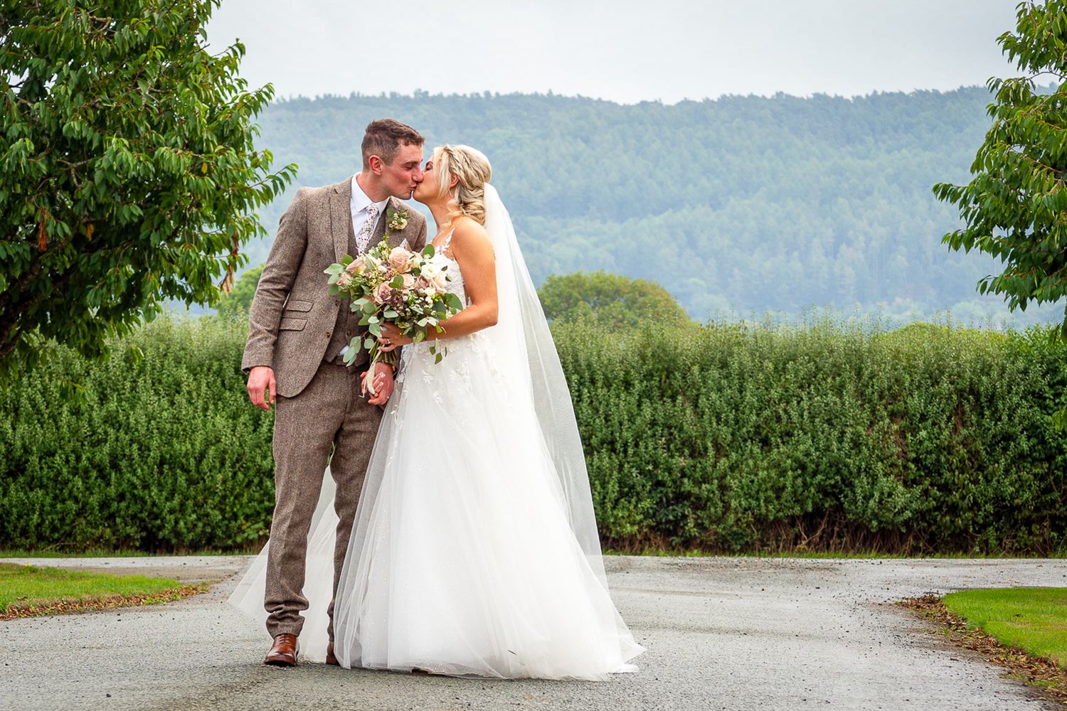 Bride and groom share a romantic kiss outdoors on their wedding day, surrounded by lush greenery and rolling hills.
