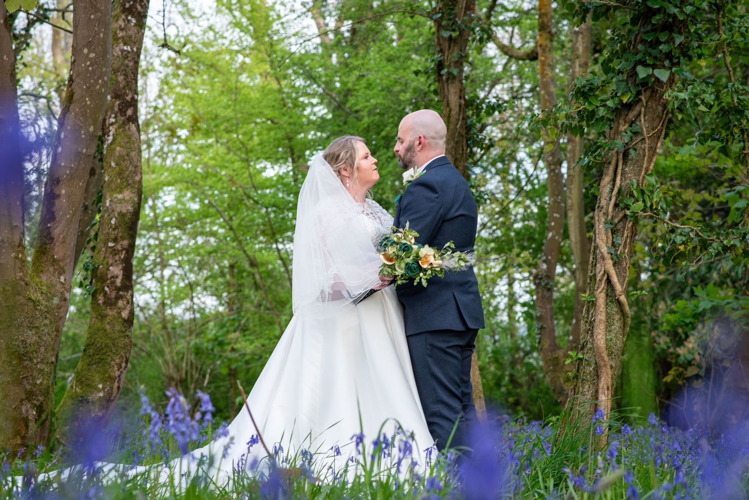 Bride and groom share a romantic moment in a bluebell woodland, bride in white gown holding colorful bouquet.