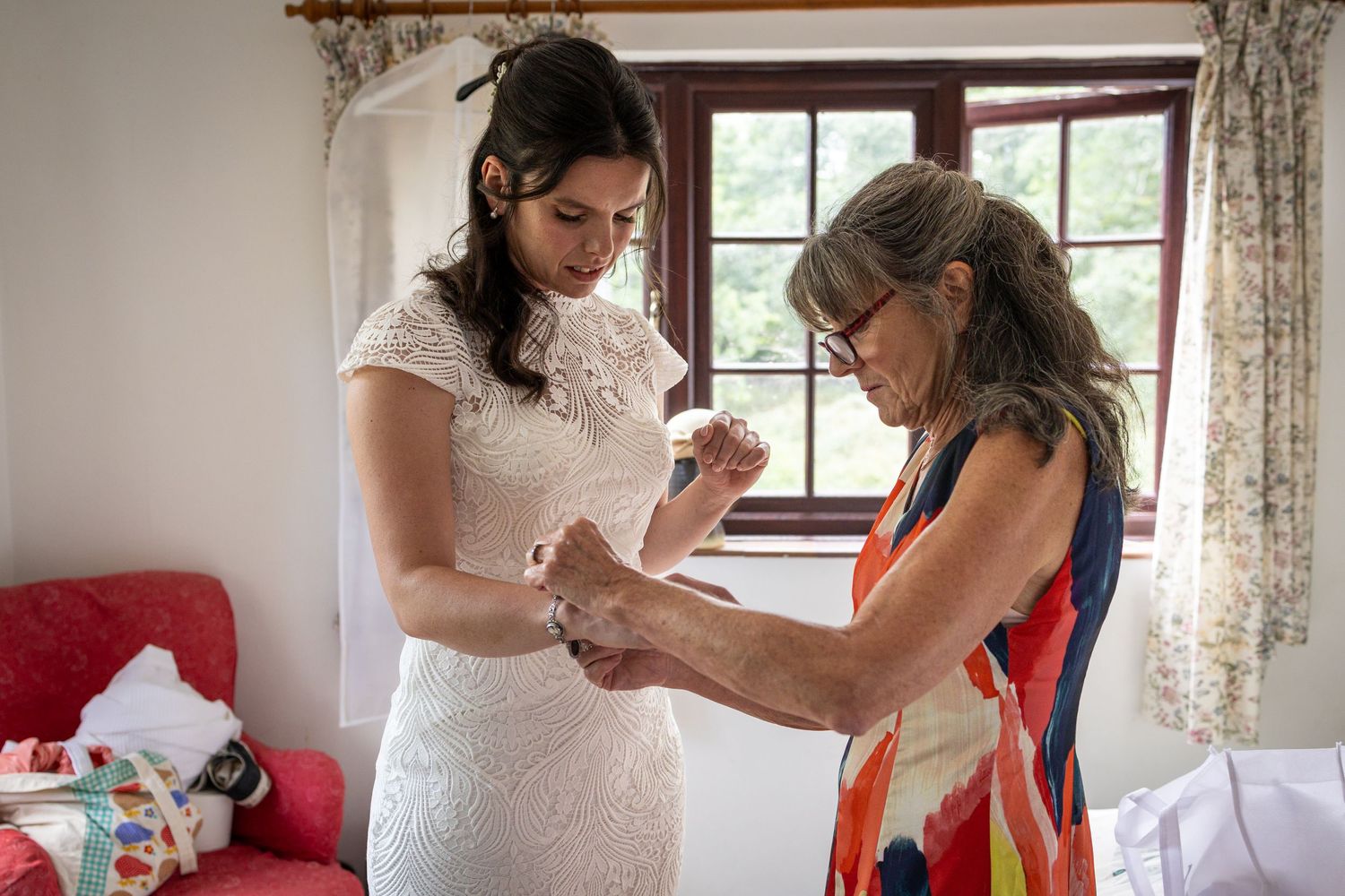 Bride in white lace dress getting help with bracelet from older woman in colorful dress before wedding ceremony.