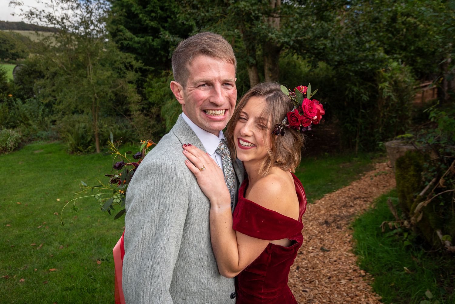Smiling couple embracing in a garden, man in grey suit and woman in red velvet off-shoulder dress with floral hair accessory.