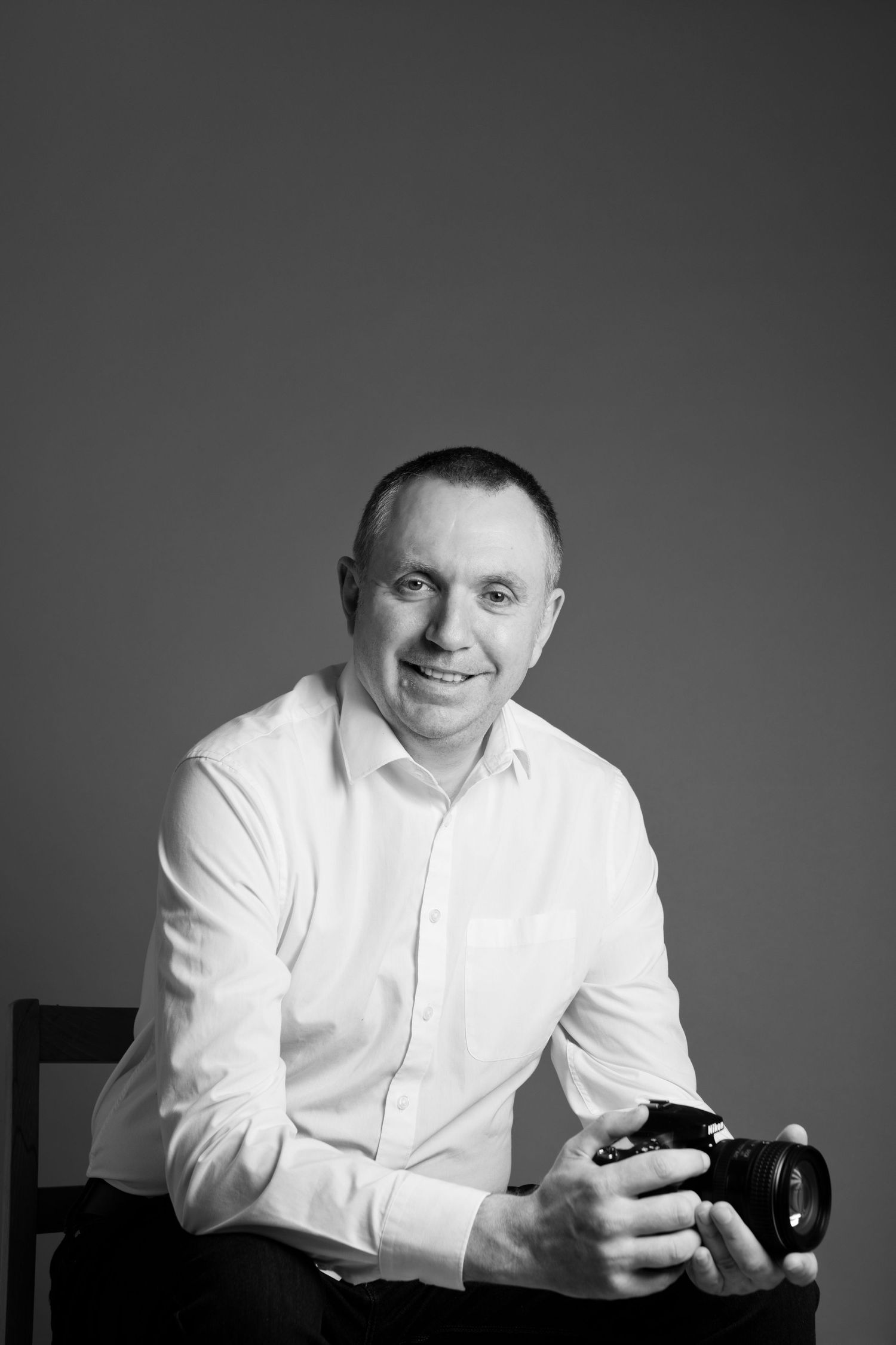 Smiling male photographer in white shirt holding DSLR camera, black and white studio portrait.
