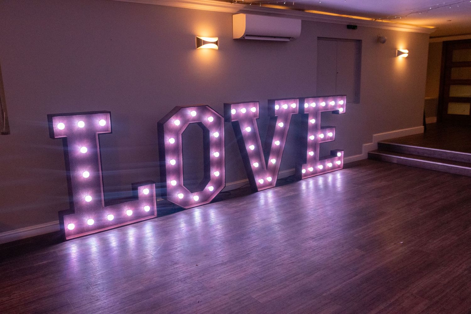 Large illuminated LOVE marquee letters glowing in purple light on wooden floor in an empty room.