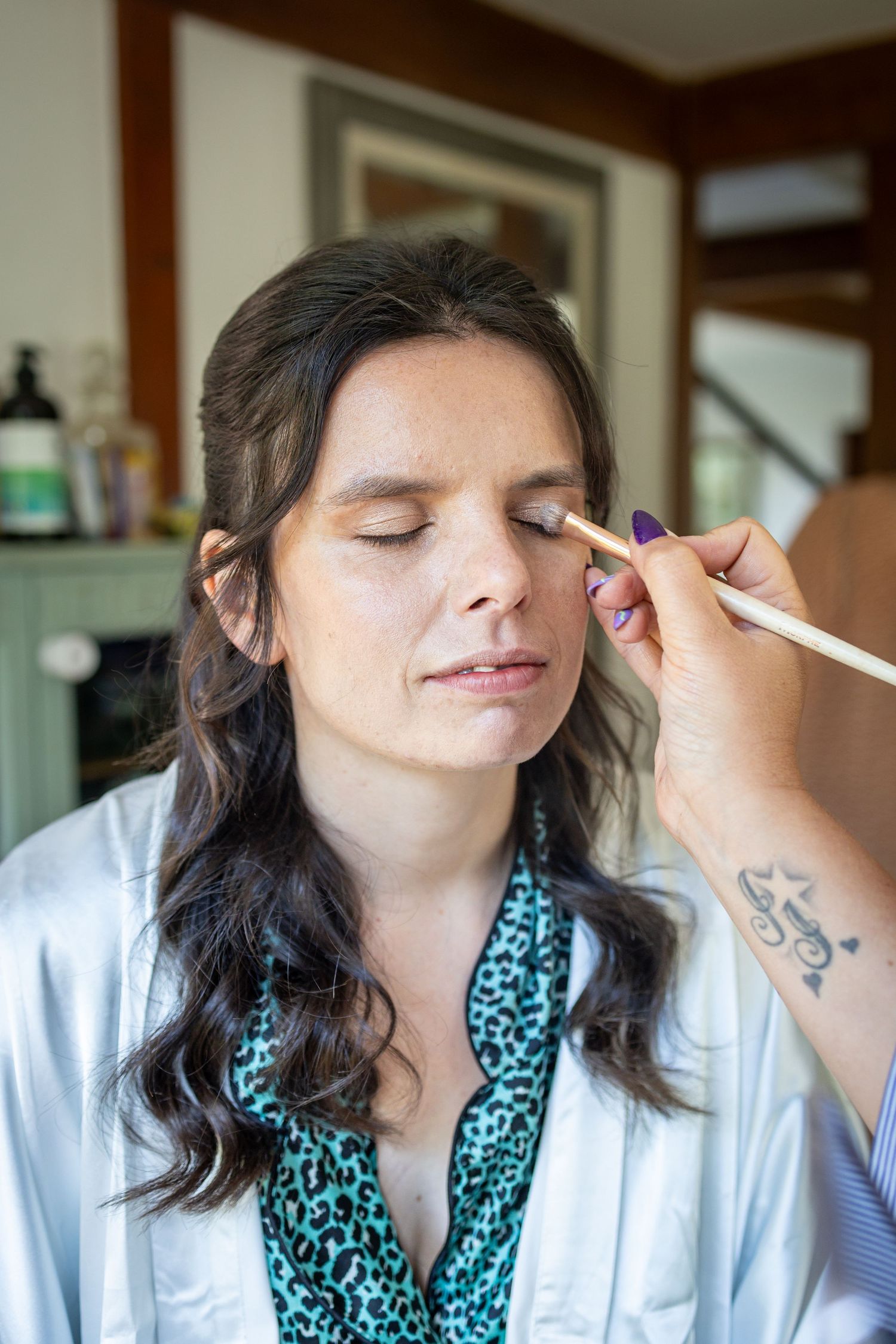 A makeup artist applies eyeshadow to a woman in a satin robe with closed eyes during bridal preparation.