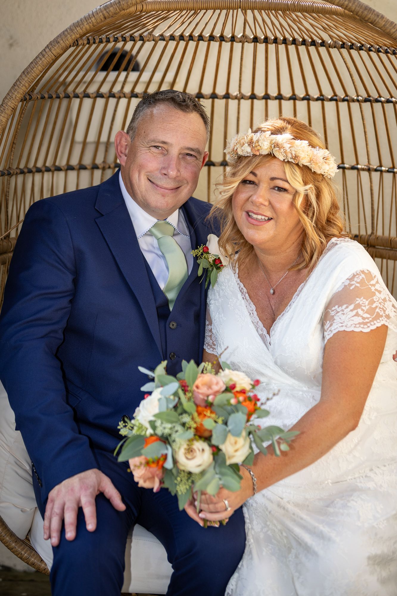 Happy couple posing in wicker egg chair, bride in lace dress with floral crown holding bouquet, groom in navy suit.