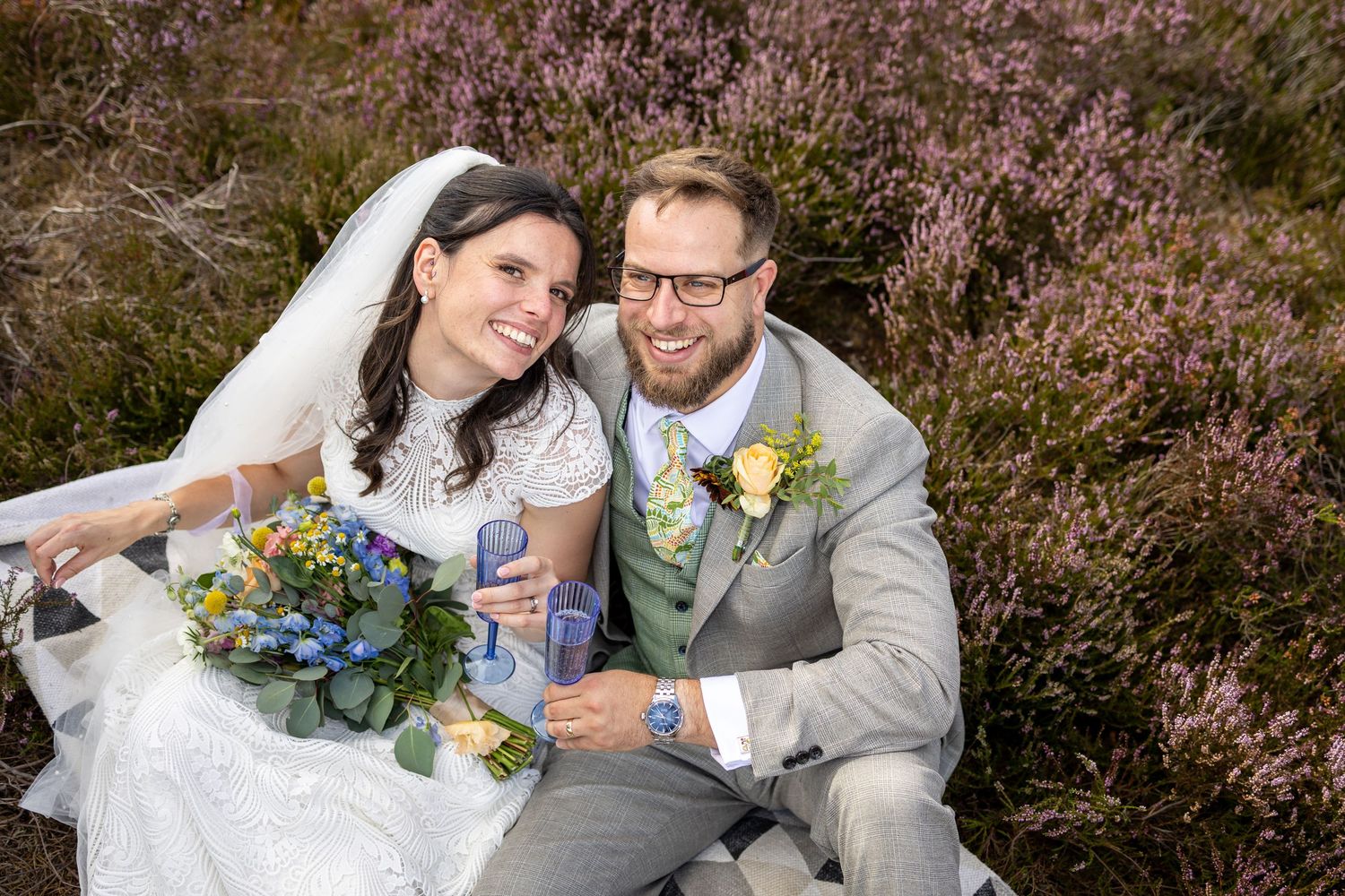 Newlyweds pose together among blooming purple heather flowers in a romantic outdoor setting.