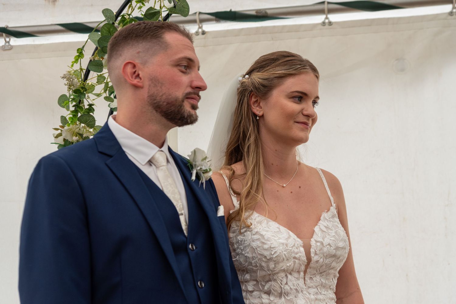 Bride and groom standing together during outdoor wedding ceremony, groom in navy suit, bride in lace dress.