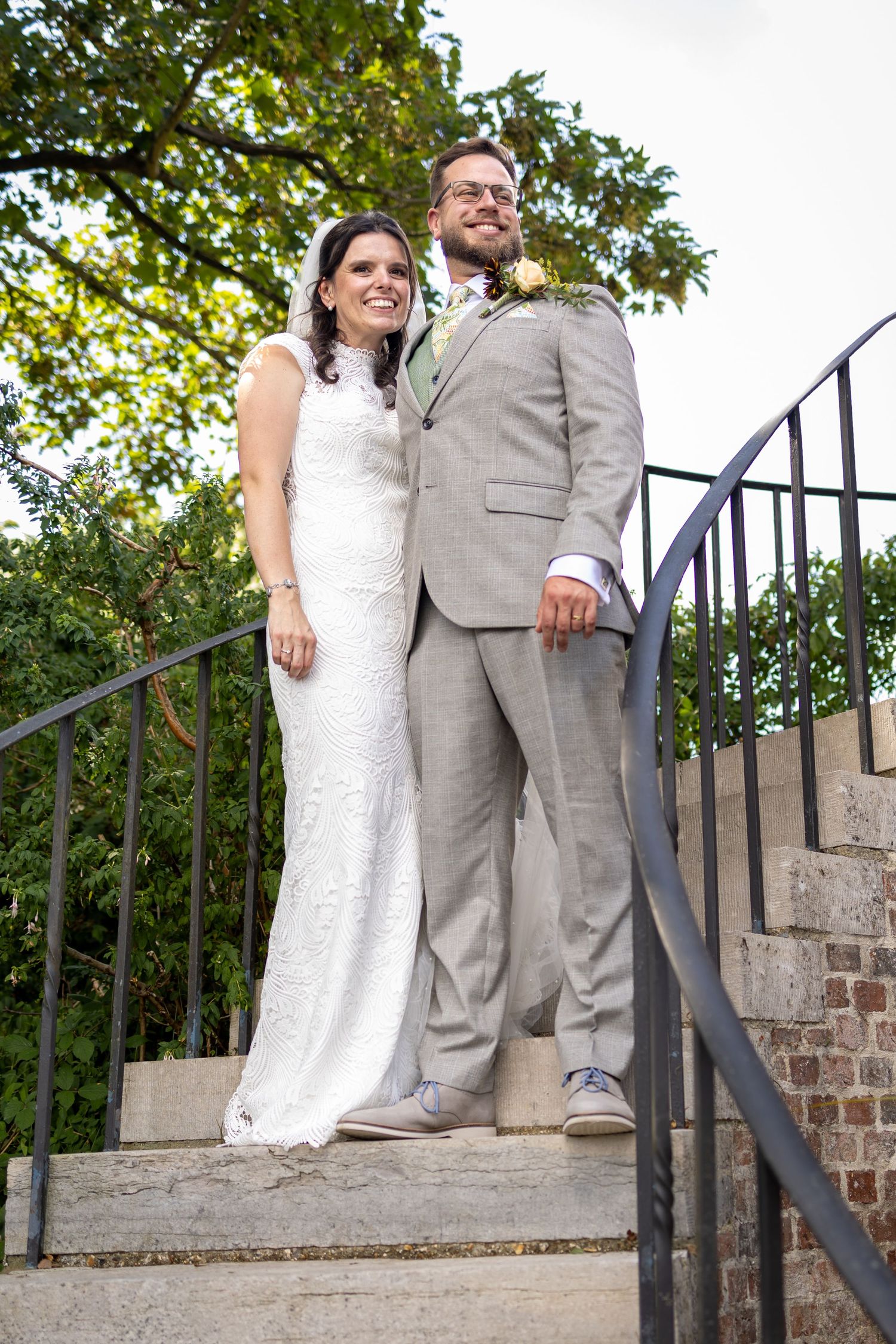 Bride in white lace gown and groom in gray suit pose together on stone steps at outdoor wedding venue.