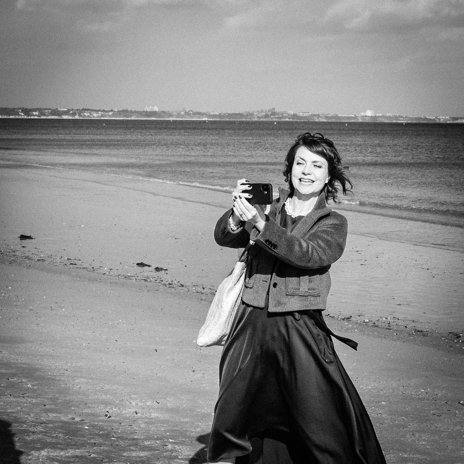 Woman smiling on beach holding camera, black and white coastal photography scene.