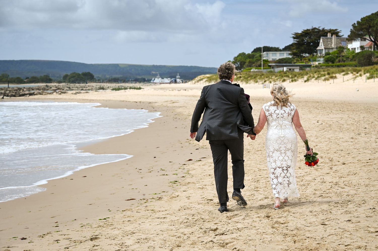 Newlywed couple walking hand in hand along a sandy beach in wedding attire, with ocean waves and green hills in background.