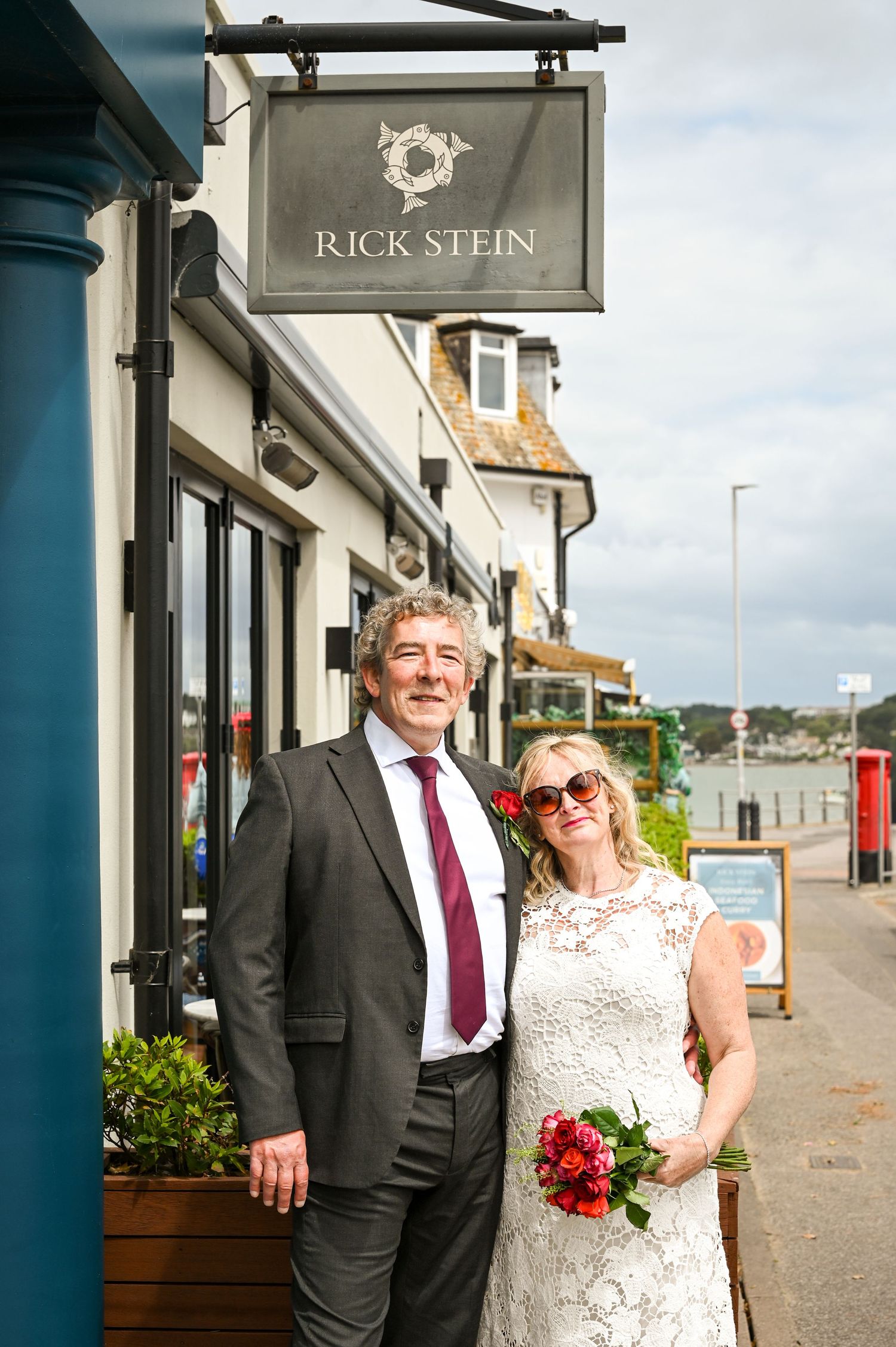 Couple in wedding attire posing outside Rick Stein restaurant, bride holding red bouquet wearing sunglasses.