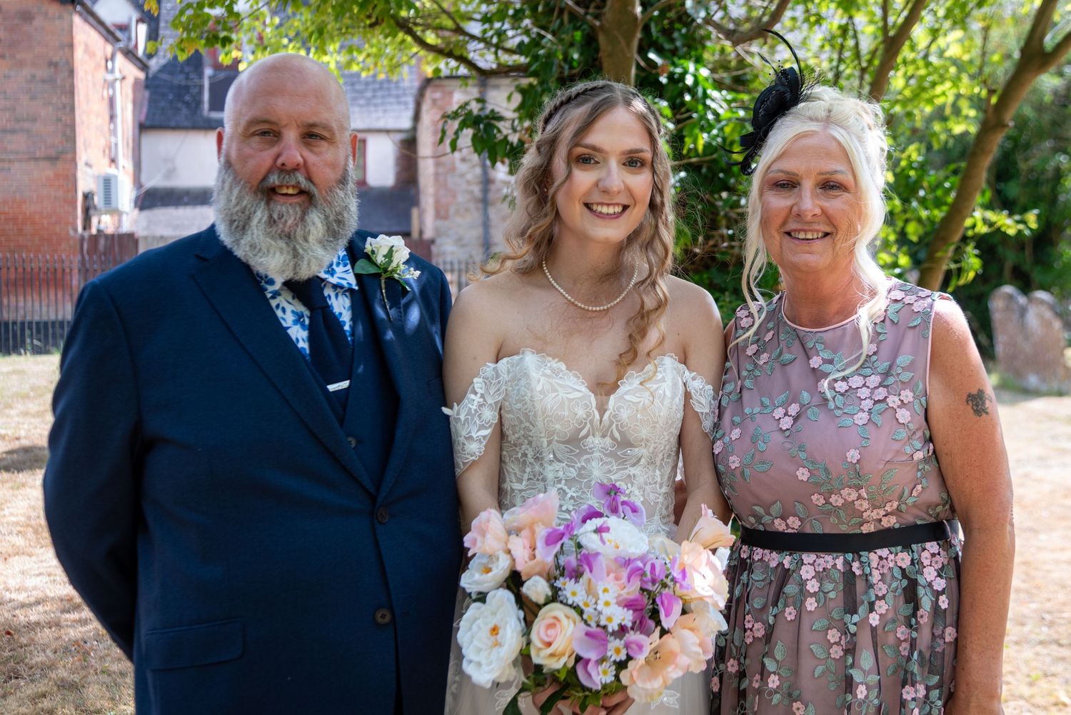 Bride poses with parents outside church, holding pastel bouquet, father in navy suit, mother in floral dress.