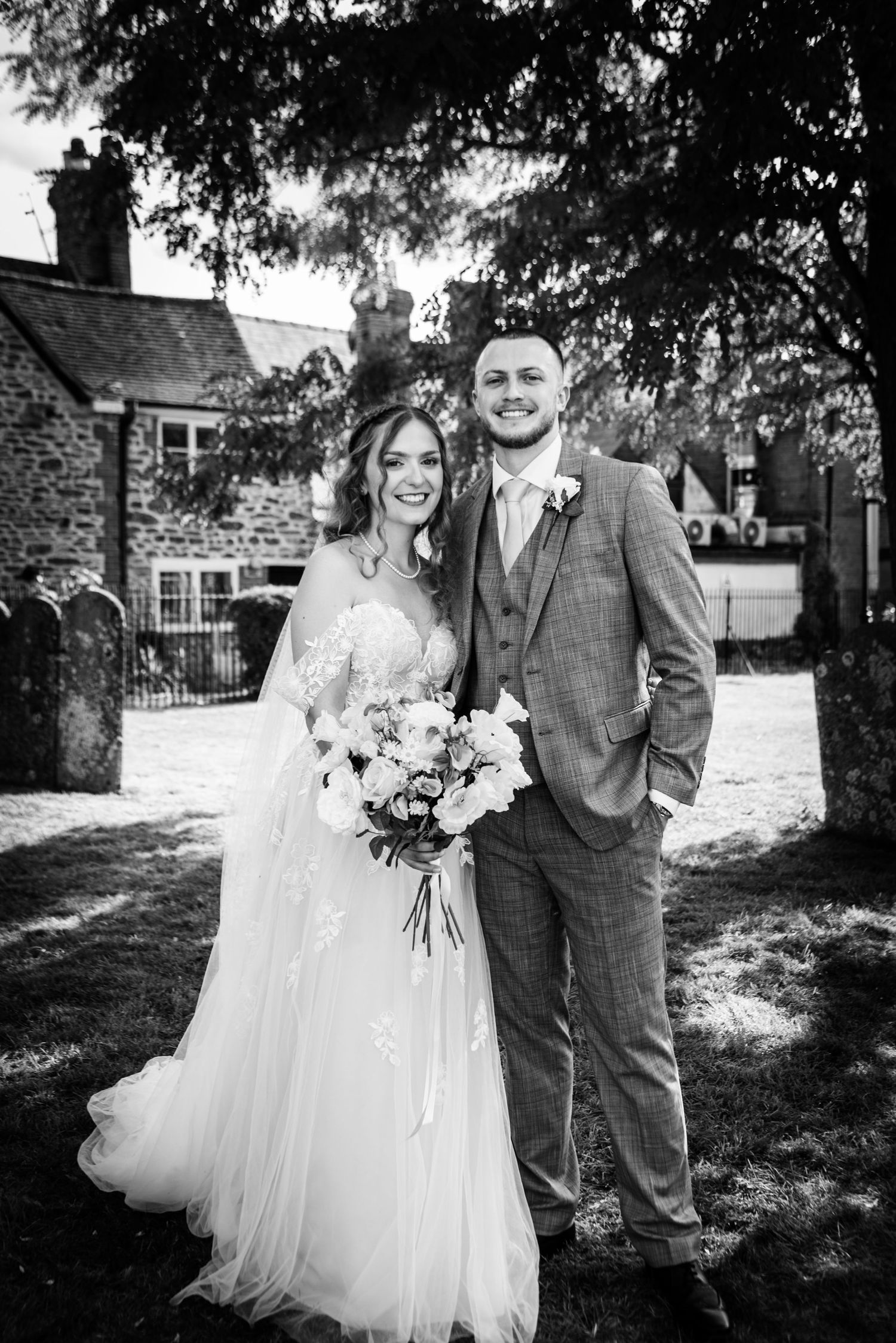 Black and white portrait of smiling bride and groom standing in a churchyard.