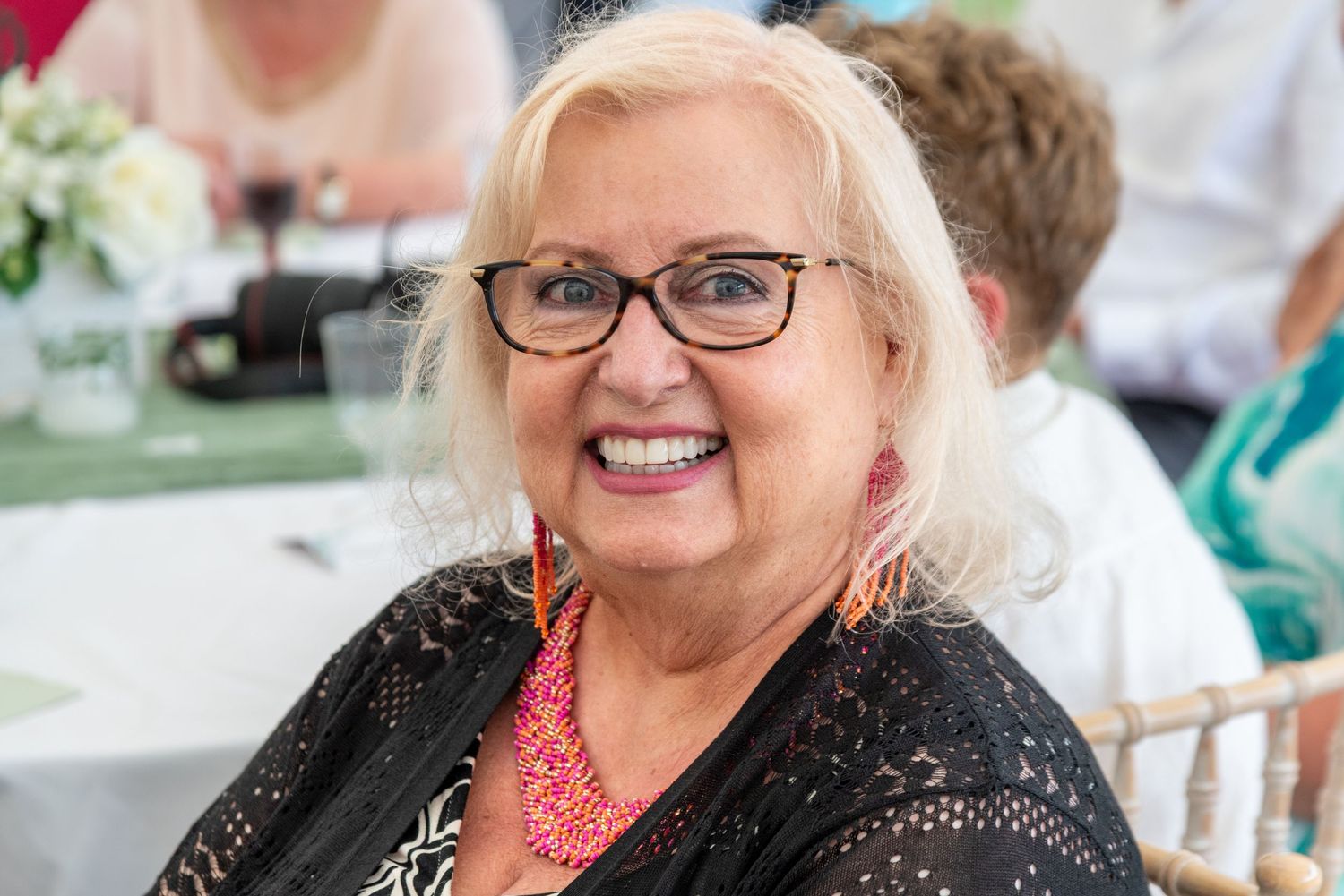 A smiling older woman with blonde hair and glasses wearing a black lace top at a wedding reception.