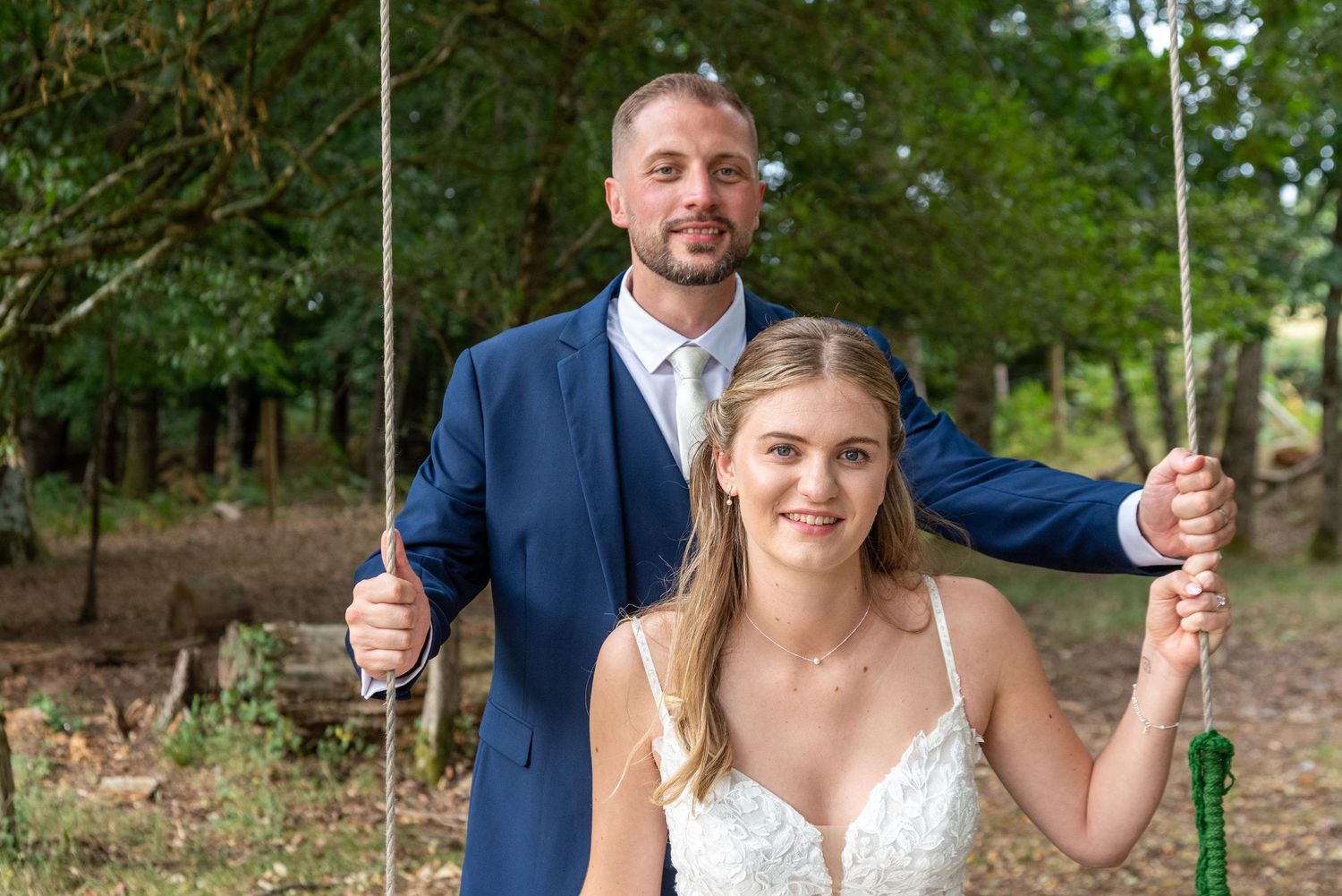 Bride sitting on a swing in a white lace dress with groom in blue suit standing behind her outdoors.