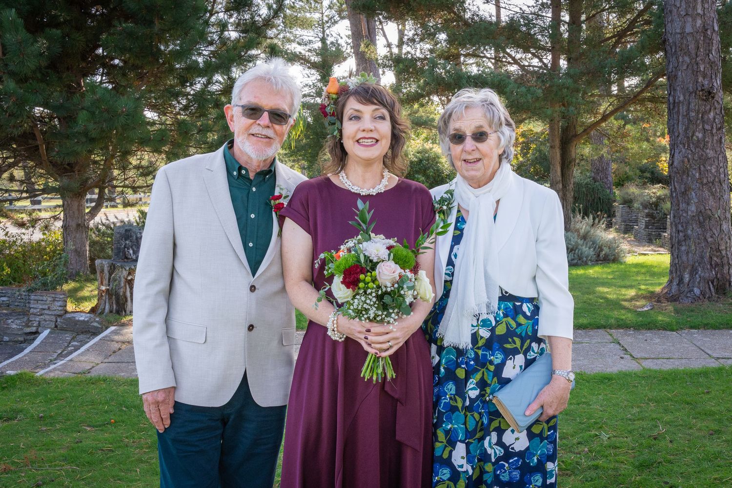 Three people pose outdoors at a wedding, woman in burgundy dress holding bouquet flanked by elderly man and woman.