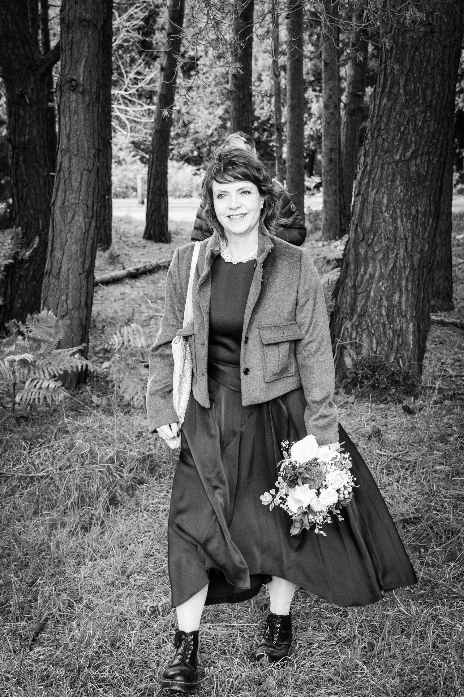 Woman smiling in forest holding bouquet, wearing jacket and black dress with boots, black and white photo.
