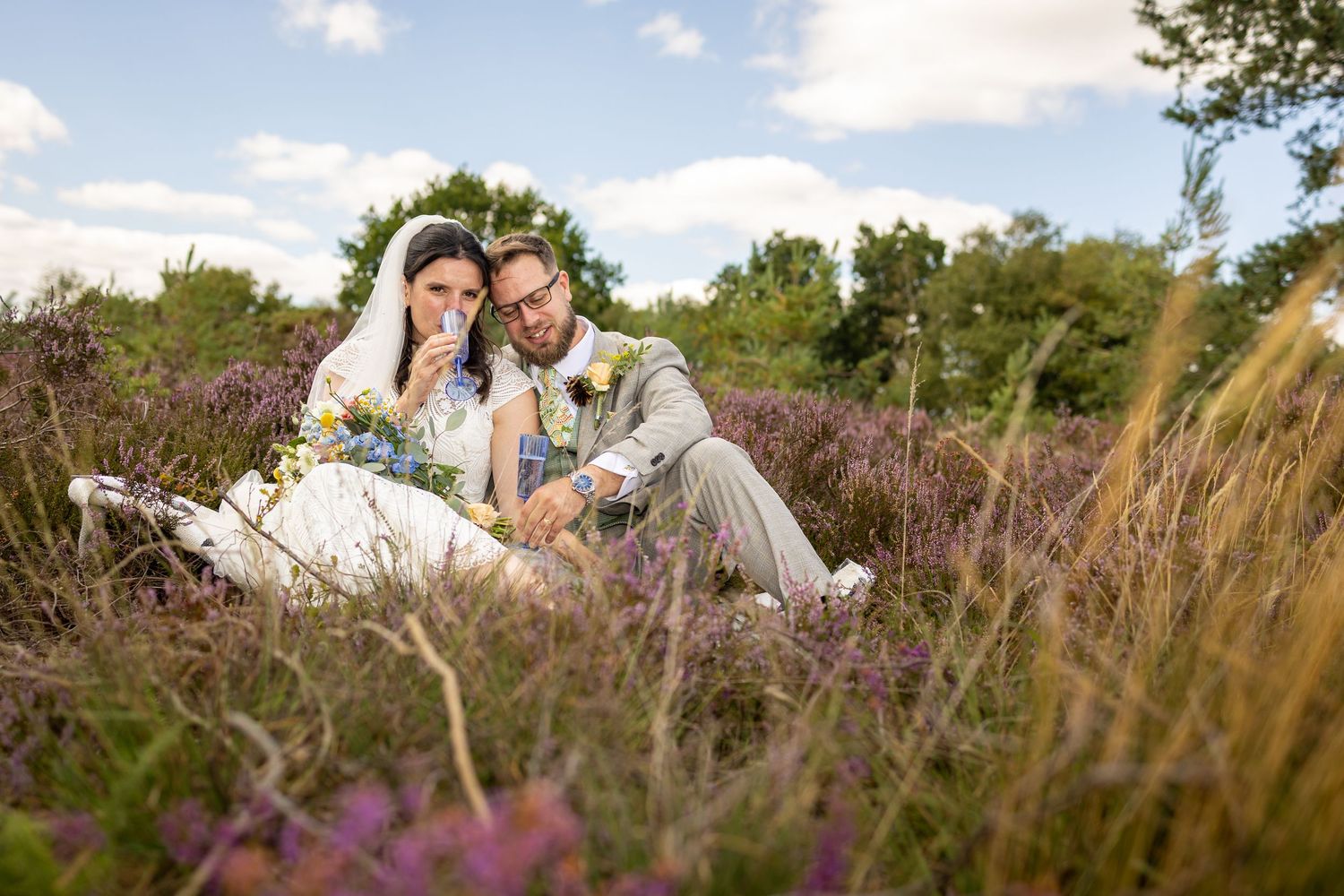 Bride and groom sitting together in purple heather field, sharing a tender moment on their wedding day under a cloudy sky.