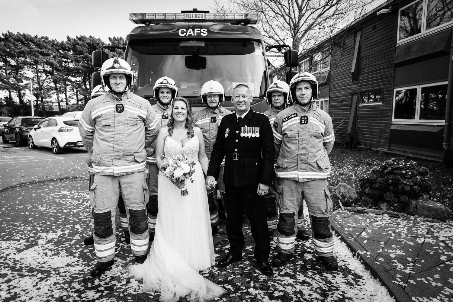Bride and groom pose with firefighters in uniform in front of a CAFS fire truck at a wedding venue.