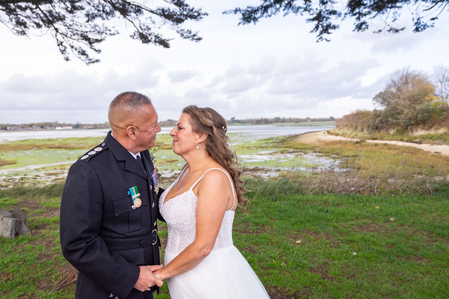 Military groom in uniform holds hands with bride in white dress outdoors by coastal scenery.
