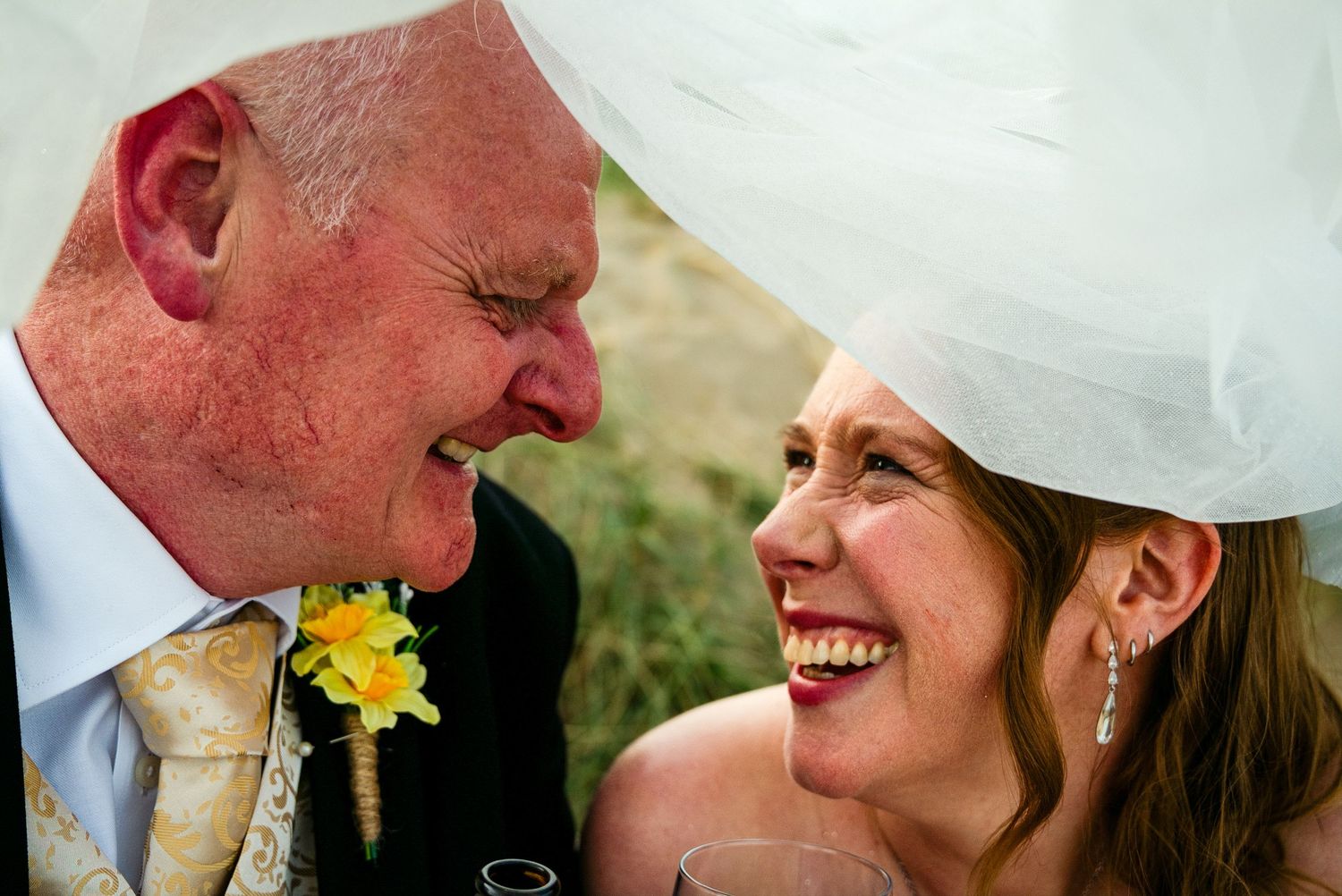A joyful bride and older man share a laugh under her wedding veil, both smiling warmly at each other.