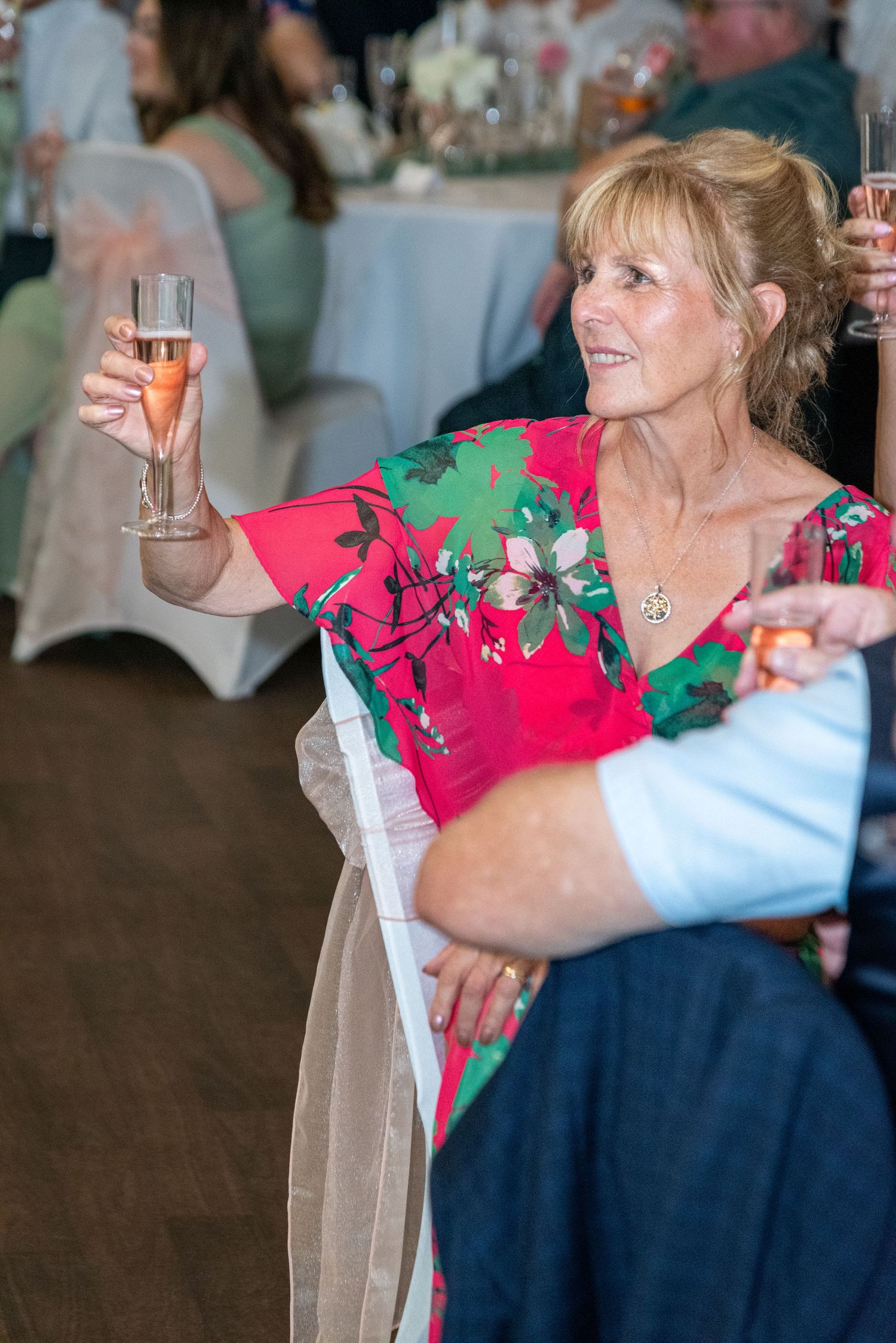 Smiling woman in red floral dress raising a champagne glass during a wedding reception toast.