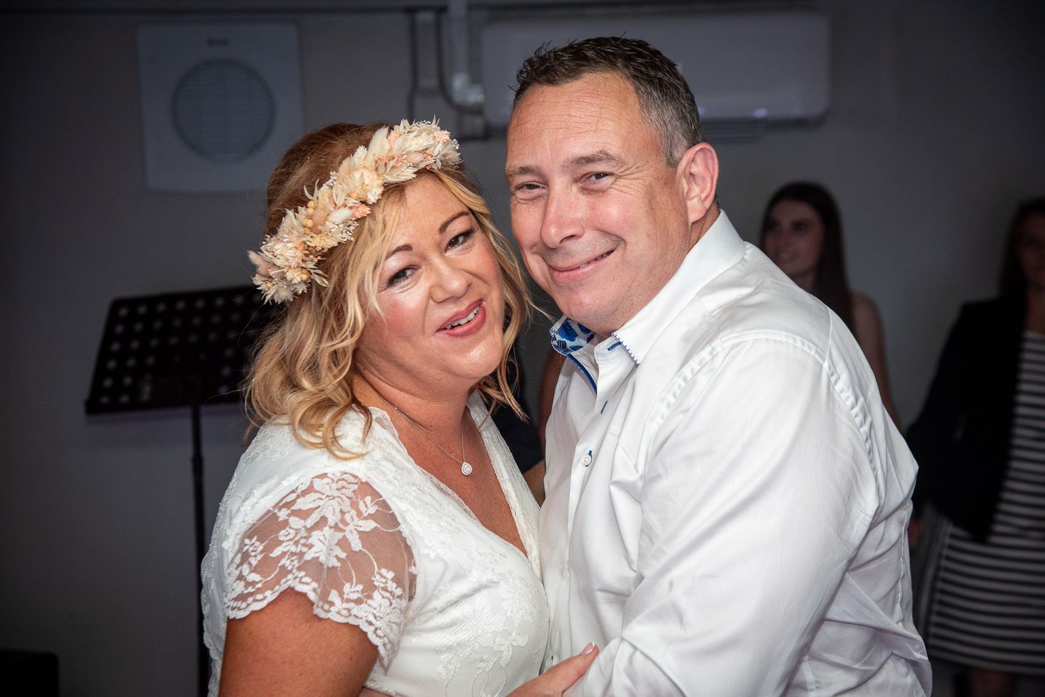Happy couple embracing and smiling at a wedding reception, bride wearing lace dress and floral crown.
