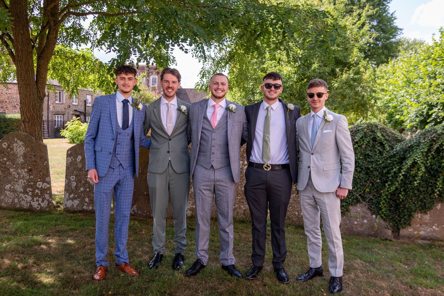 Five men in suits posing outdoors at a wedding, smiling under green trees on a sunny day.