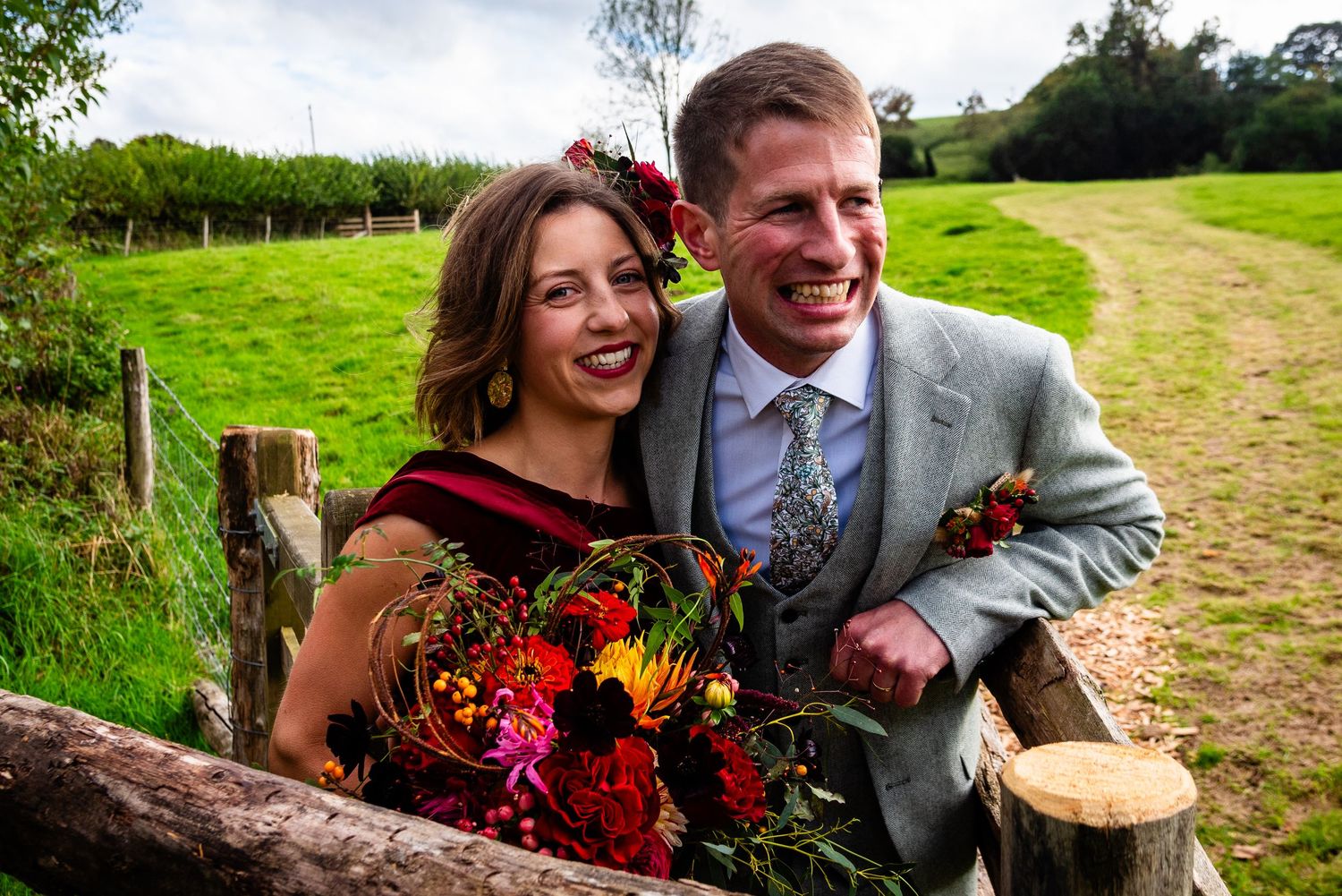 Smiling couple in wedding attire pose by rustic wooden fence with colorful bridal bouquet in lush green countryside.