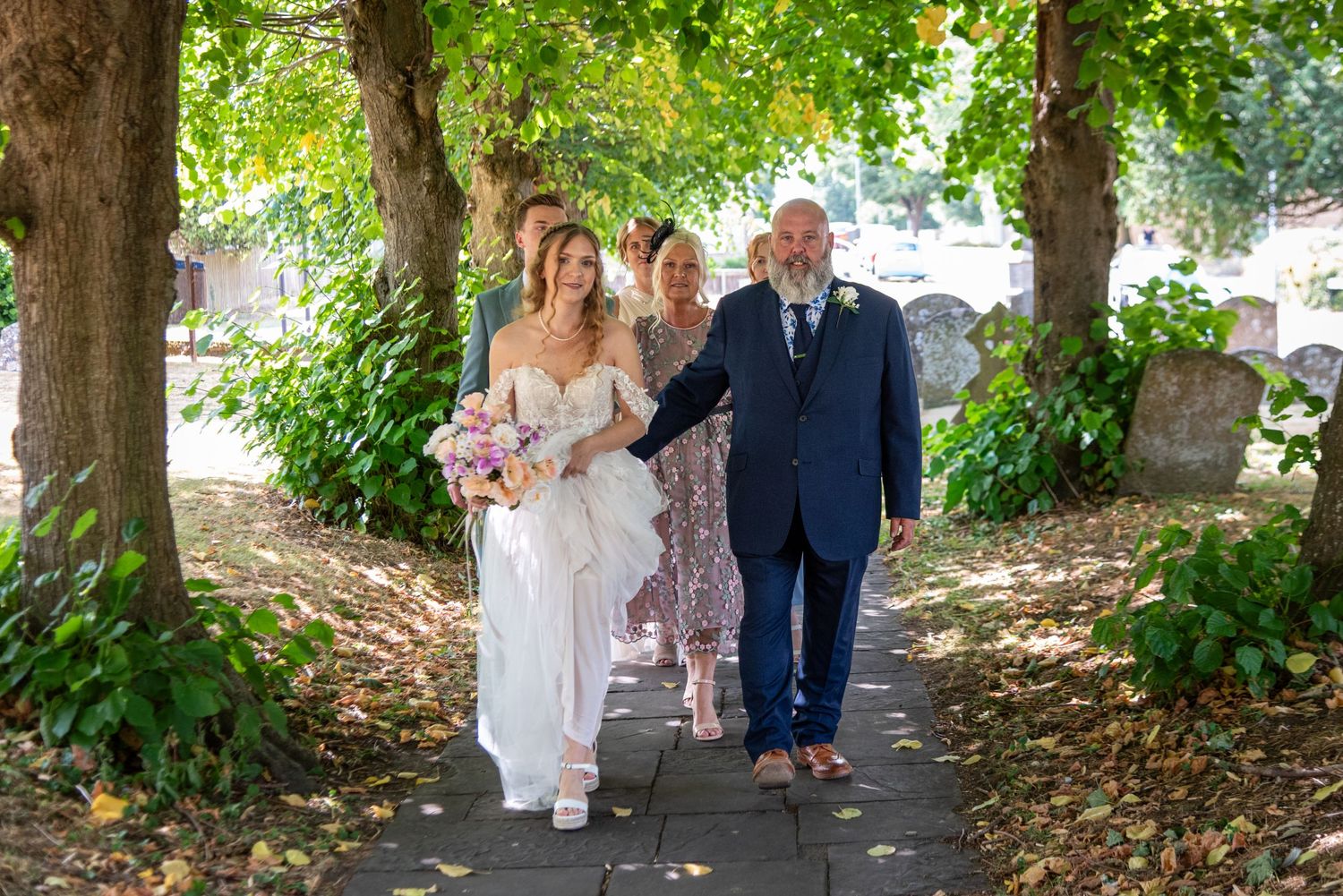 Bride in white gown walks with family along a tree-lined church path holding a pink bouquet on her wedding day.