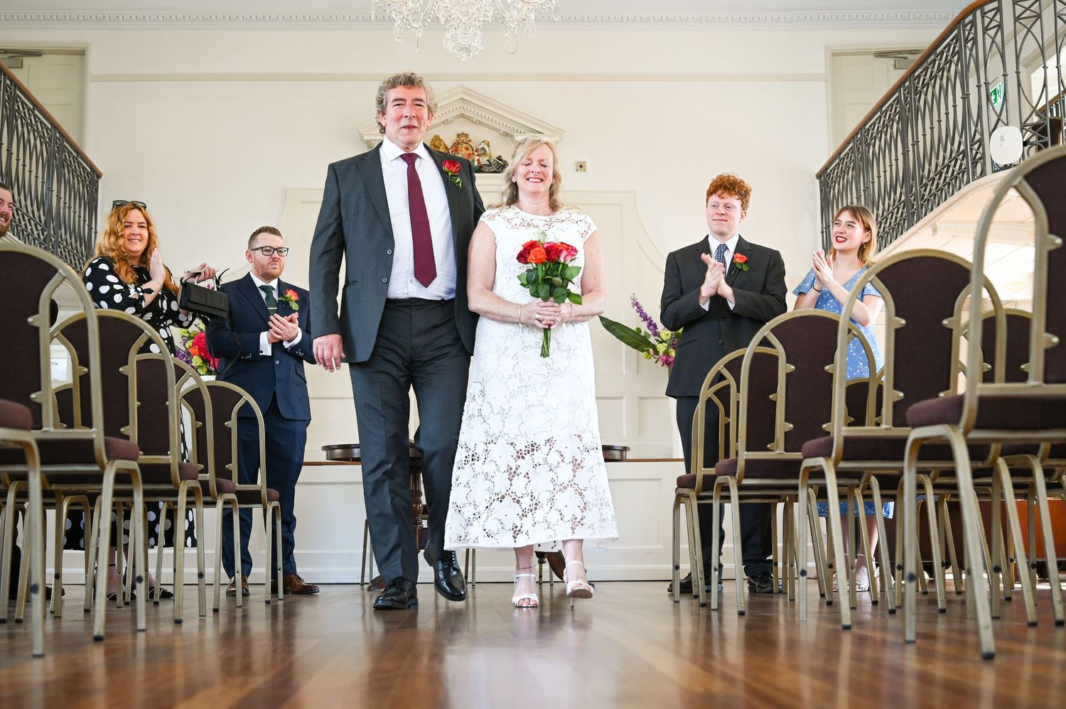 Couple walks down aisle at wedding ceremony in elegant hall, guests applauding in background.